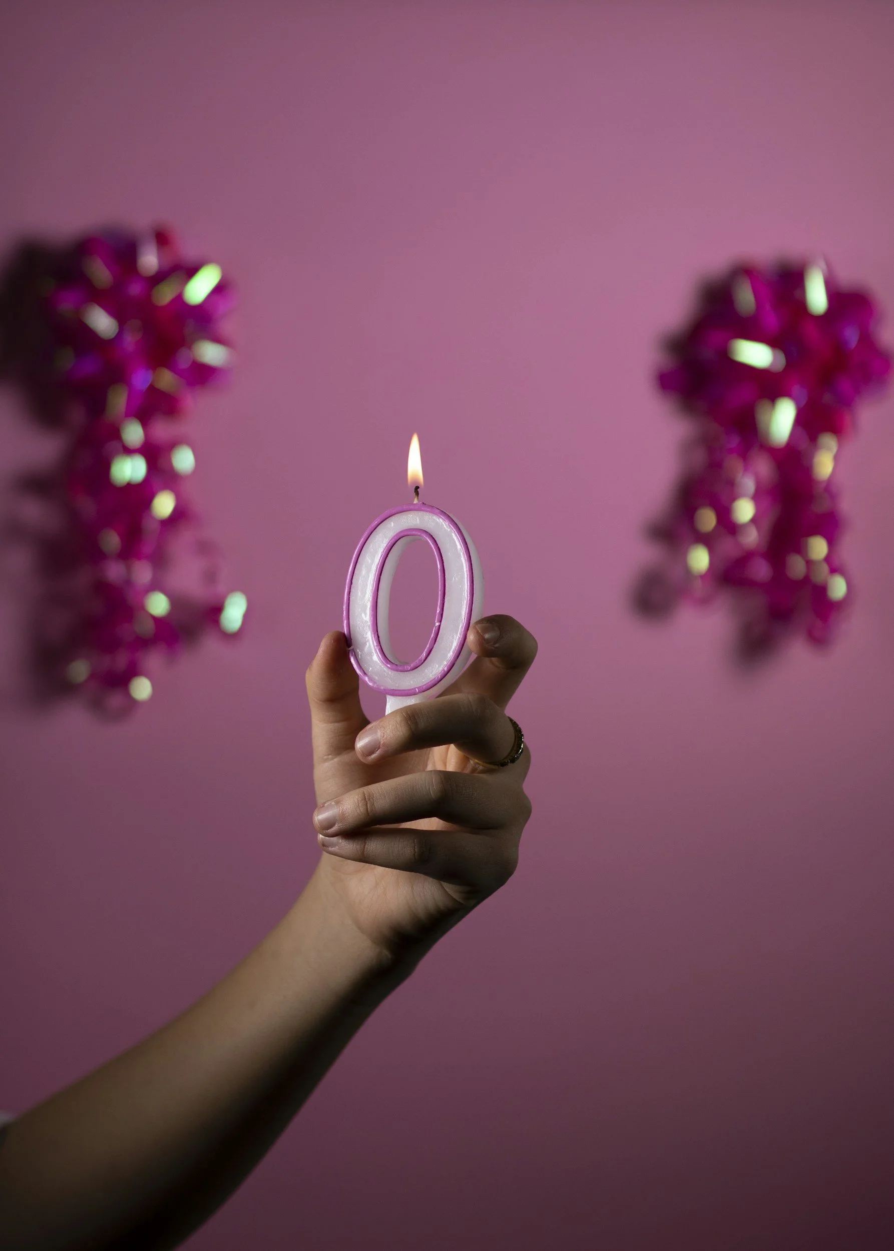 A hand holding a pink candle shaped like the number 0 with a small flame, against a pink background with two blurred pink decorative objects on either side.