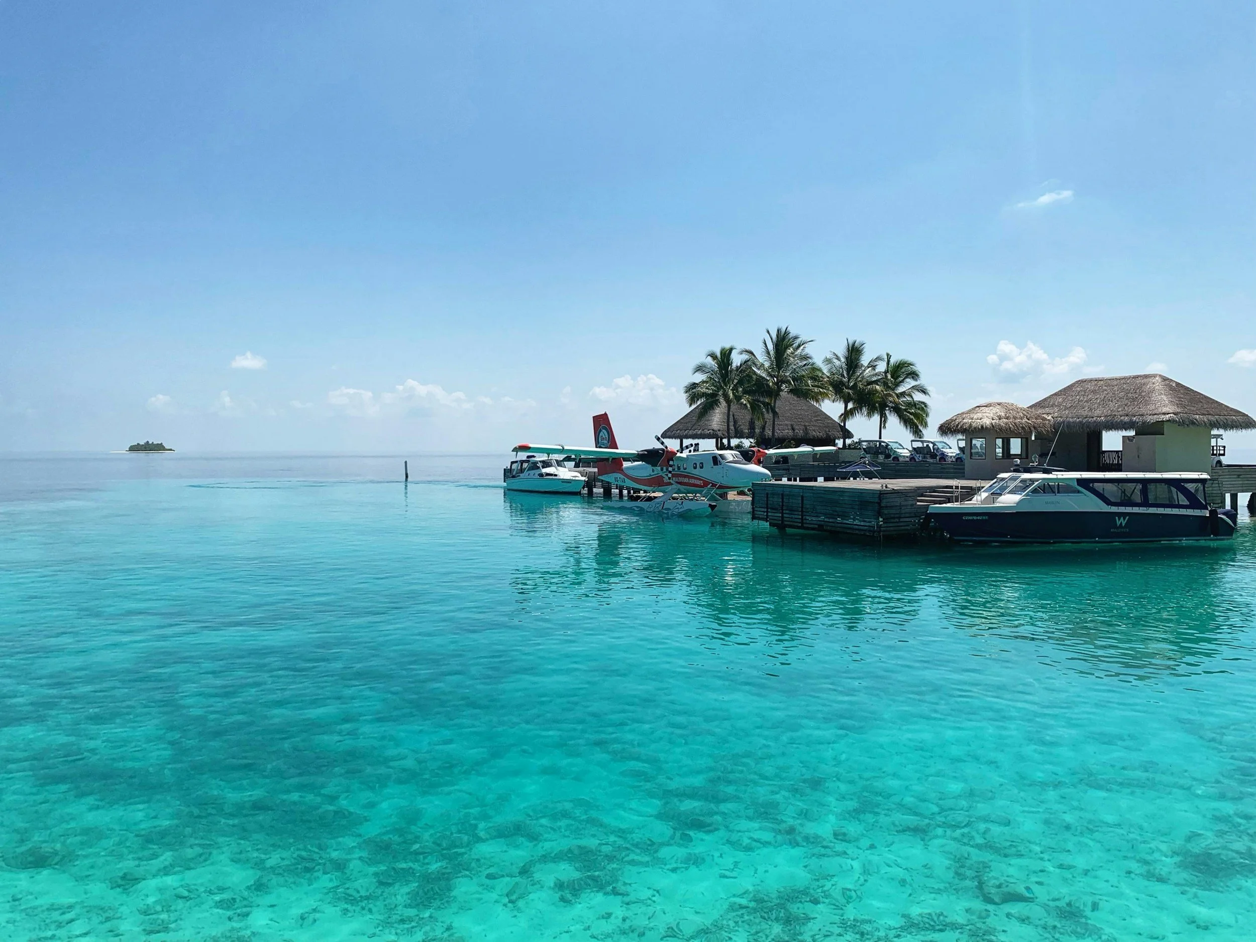 Une vue d'un quai avec des bateaux, des palmiers, et des huttes en tôle à un bord d'une mer turquoise claire, avec un ciel bleu clair et quelques petits nuages.