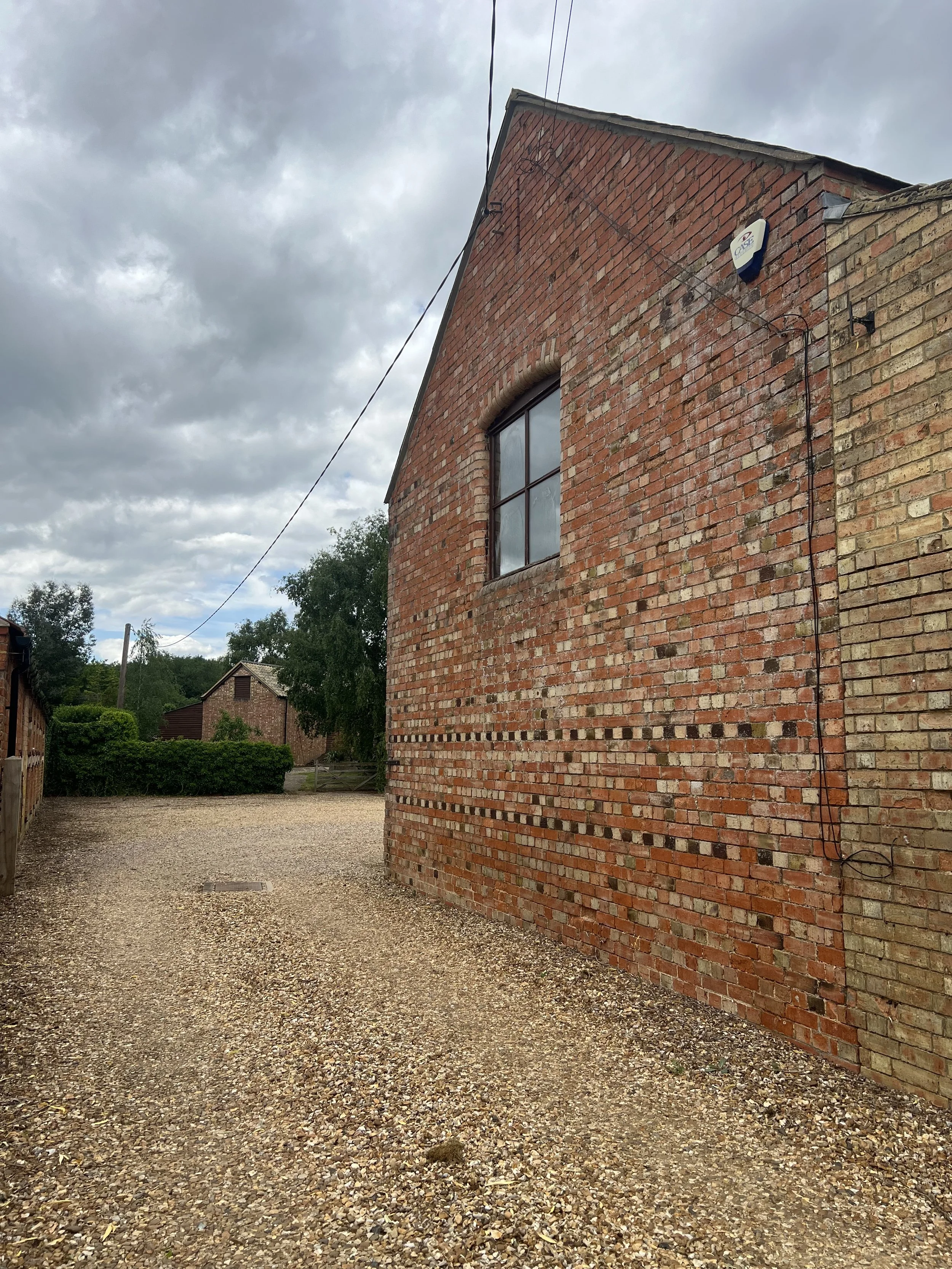 A red brick building with a window on the side, an overcast sky, and a gravel pathway along the building.