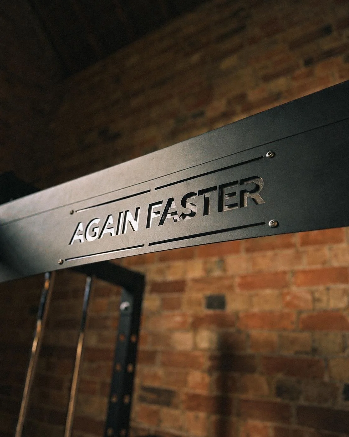 Close-up of a metal sign with the words "AGAIN FASTER" cut out, mounted on a black metal frame against a brick wall.