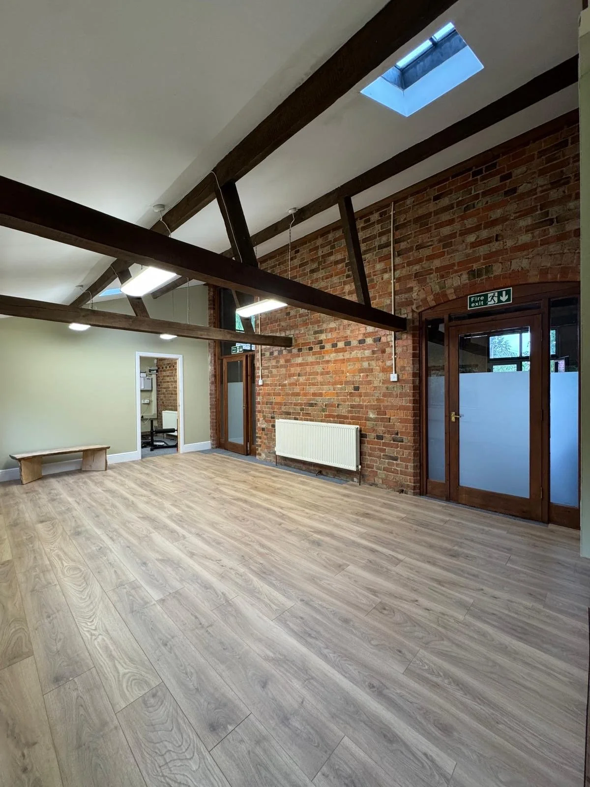 An empty room with wooden flooring, a brick wall, exposed ceiling beams, and large glass doors with a fire exit sign above.