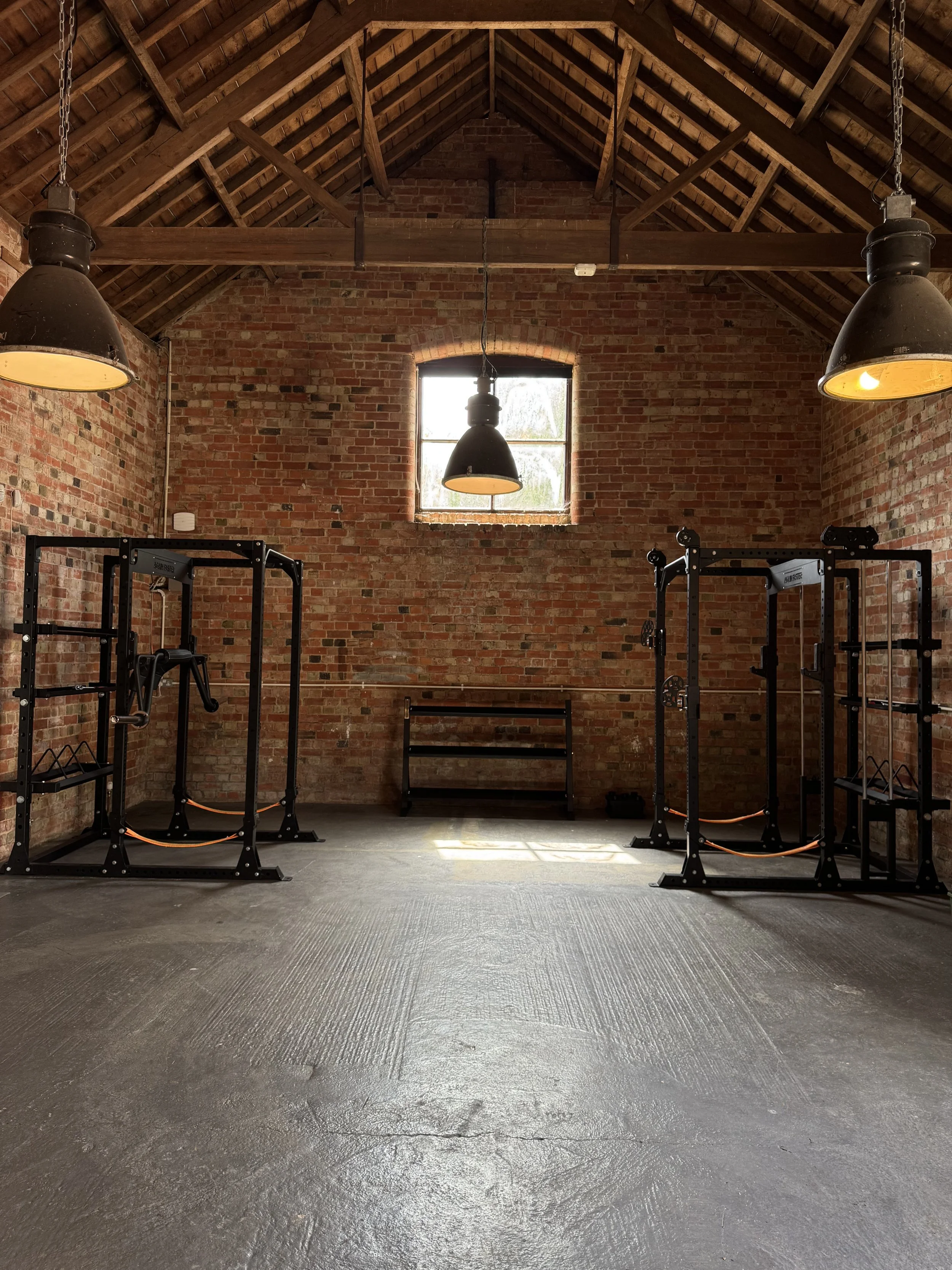 An indoor gym or workout space with brick walls, a high ceiling with exposed wooden beams, large industrial hanging lights, and black gym equipment such as squat racks and benches. There is a small window on the back wall allowing natural light to enter.