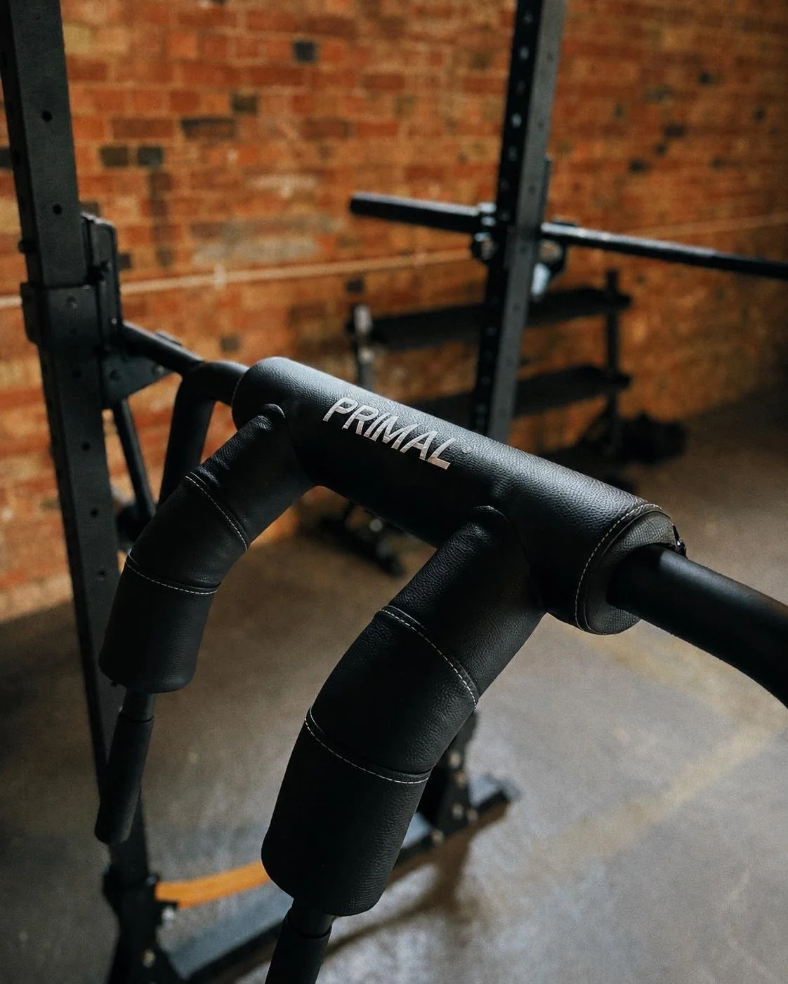 Close-up of a black PRIMAL kettlebell handle in front of a brick wall and gym rack.