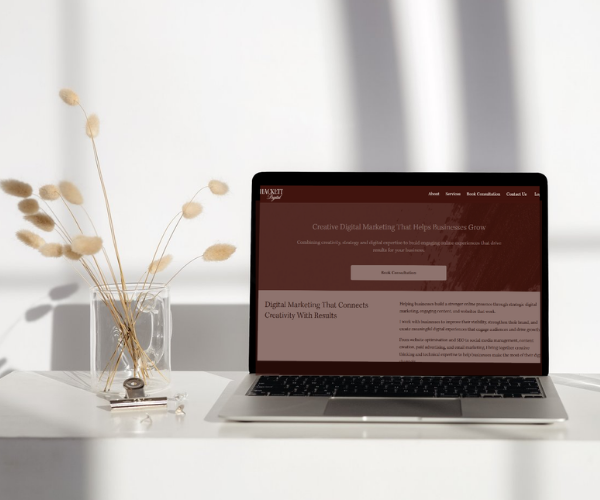 A laptop displaying a Hackett Digital digital marketing website on a white desk, with a glass vase holding dried beige flowers to the left.