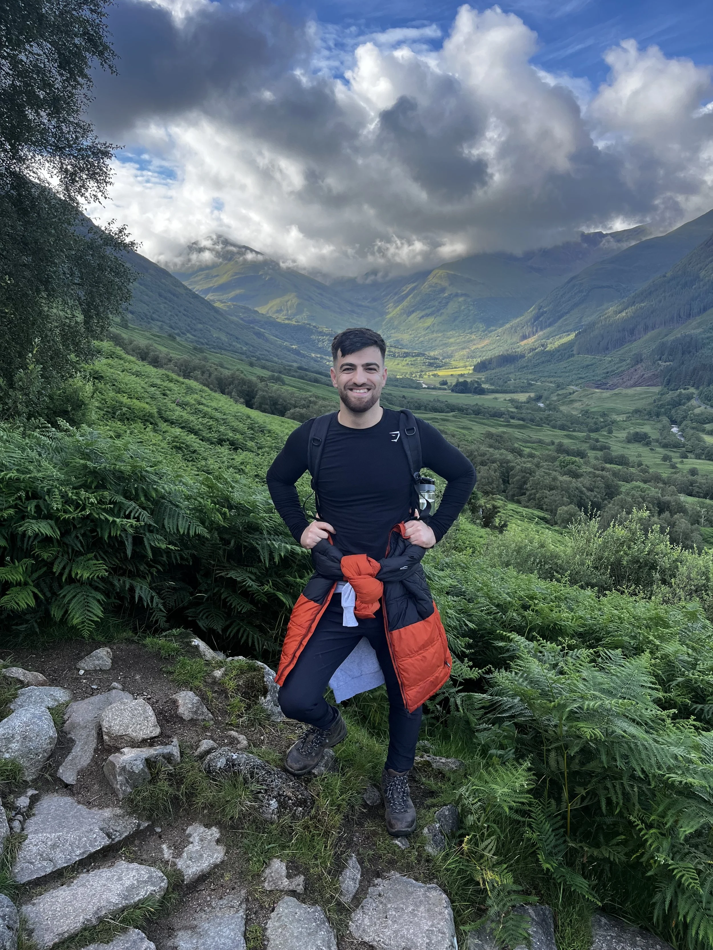 A man with short dark hair and a beard, smiling and standing outdoors in a mountainous landscape. He is wearing a black long-sleeved shirt, hiking boots, and has a red and black jacket tied around his waist. The background features green rolling hills, mountain slopes, and a sky filled with clouds.