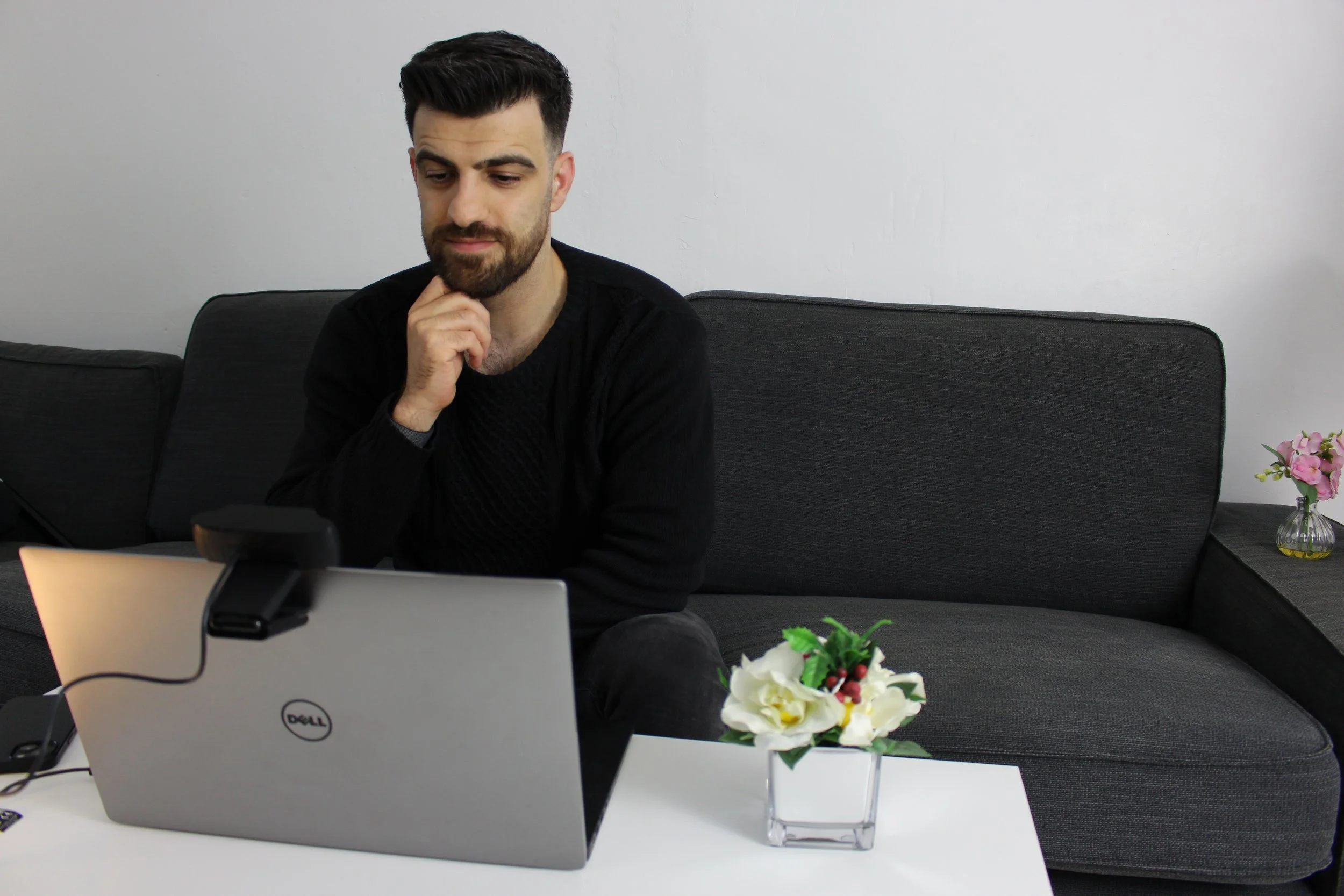 A young man with dark hair and a beard sitting on a gray sofa, looking at a laptop in front of him, with a white wall behind him. There is a small bouquet of pink and white flowers on the white table in front of him.