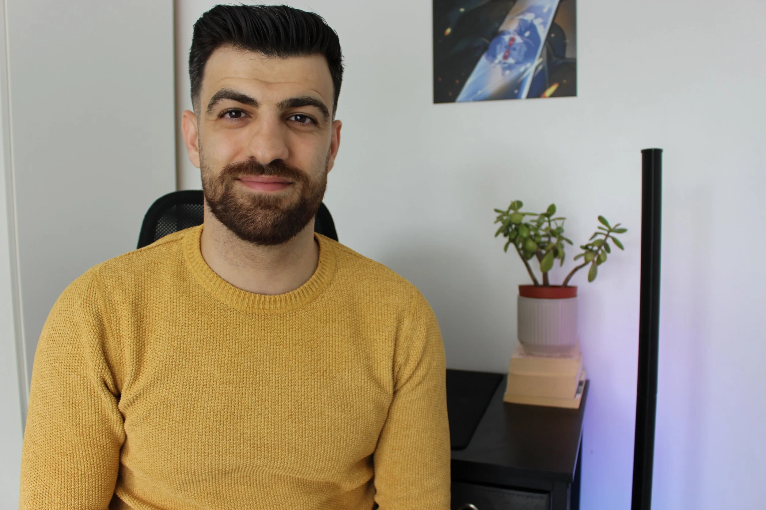 A man with dark hair and a beard wearing a yellow sweater, sitting in a black chair in an office with a white wall, a plant on a table, and a colorful abstract art piece on the wall.