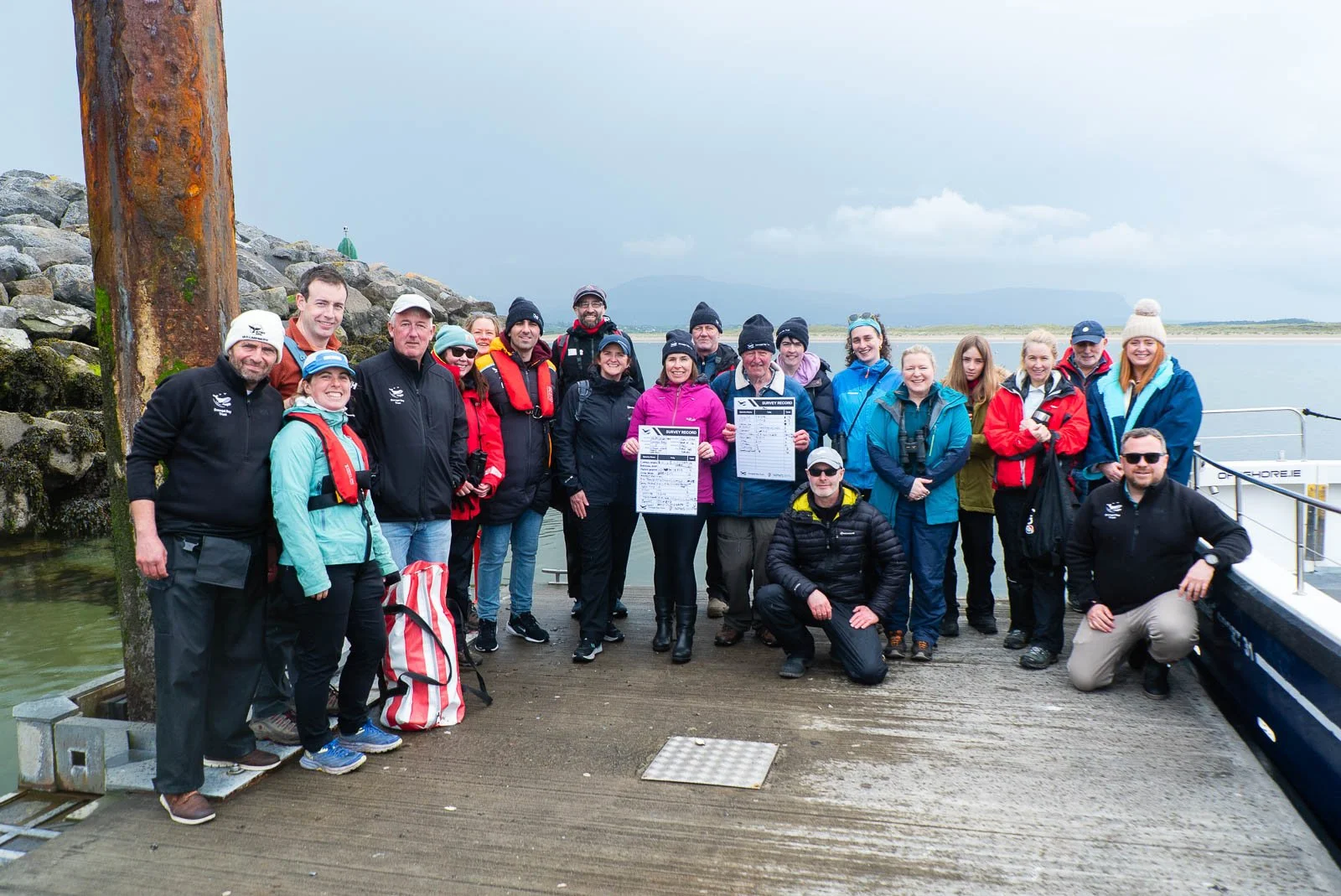 Group of people gathered on a dock near water, holding survey records, dressed in outdoor and waterproof clothing, with mountains and cloudy sky in the background.