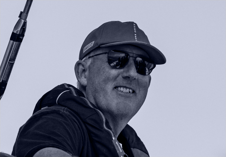 Man in sunglasses and baseball cap smiling outdoors, carrying a fishing pole.
