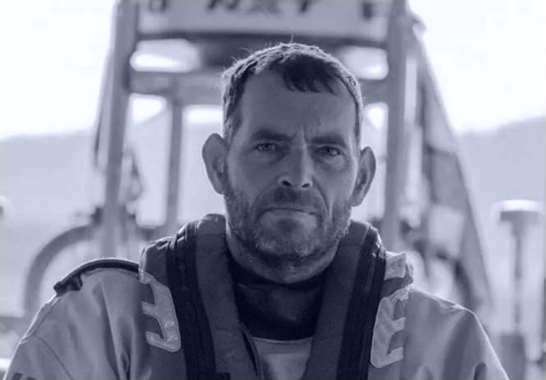 Black and white photo of a man in outdoor gear with a serious expression, sitting in front of a vehicle.