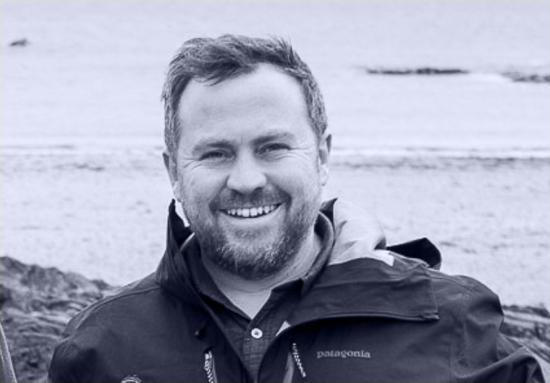 A smiling man with short hair and a beard standing outdoors near a body of water, wearing a Patagonia jacket.