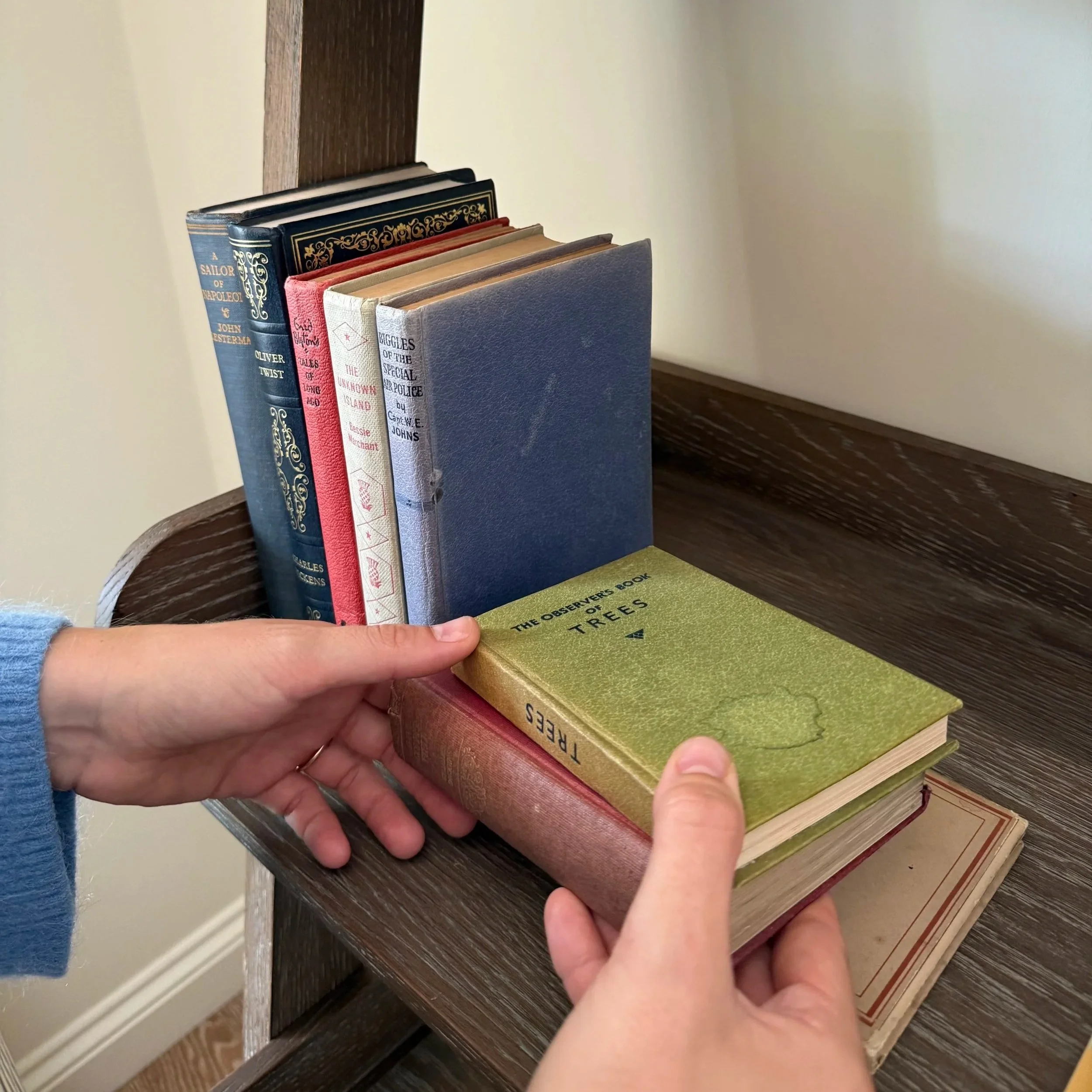 A person, wearing a blue sweater, holding a book with a yellow-green cover titled 'The Observers Book of Trees,' standing next to a shelf with several other books.