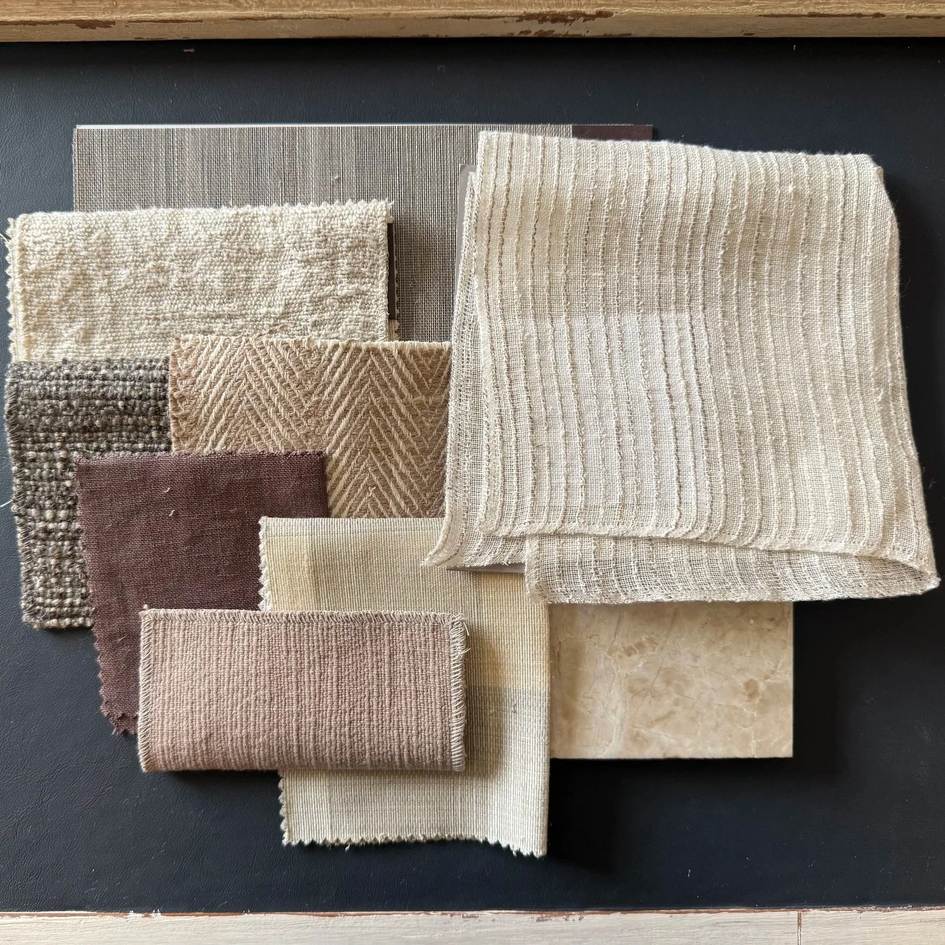 Six folded fabric swatches with various textures and patterns placed on a black surface, with a wooden surface partially visible at the top of the image.