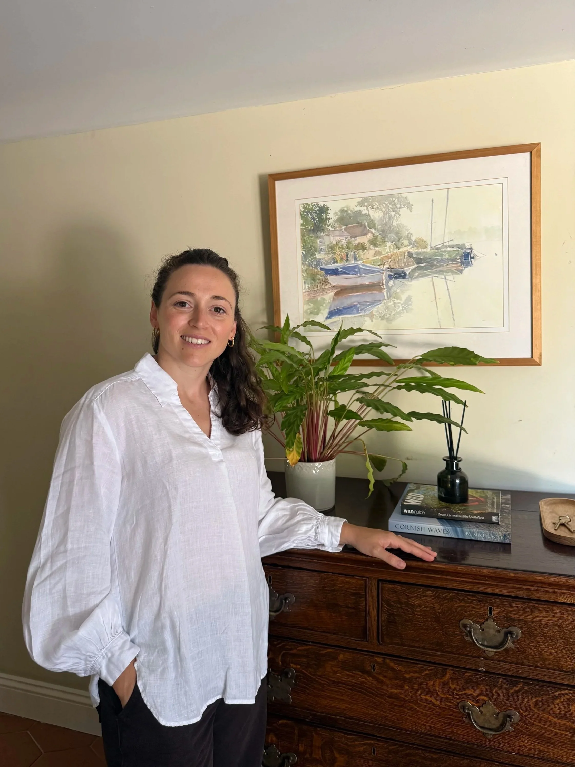 A woman with long dark hair, wearing a white shirt with rolled-up sleeves, is standing next to a wooden chest of drawers. She is smiling and has one hand resting on the chest. Behind her, on the wall, there is a framed watercolor painting of boats on water, trees, and a building. On the surface of the chest, there is a tall potted plant with green leaves, a black reed diffuser, and a stack of books titled 'WILD Guide' and 'Cornish Waves'.