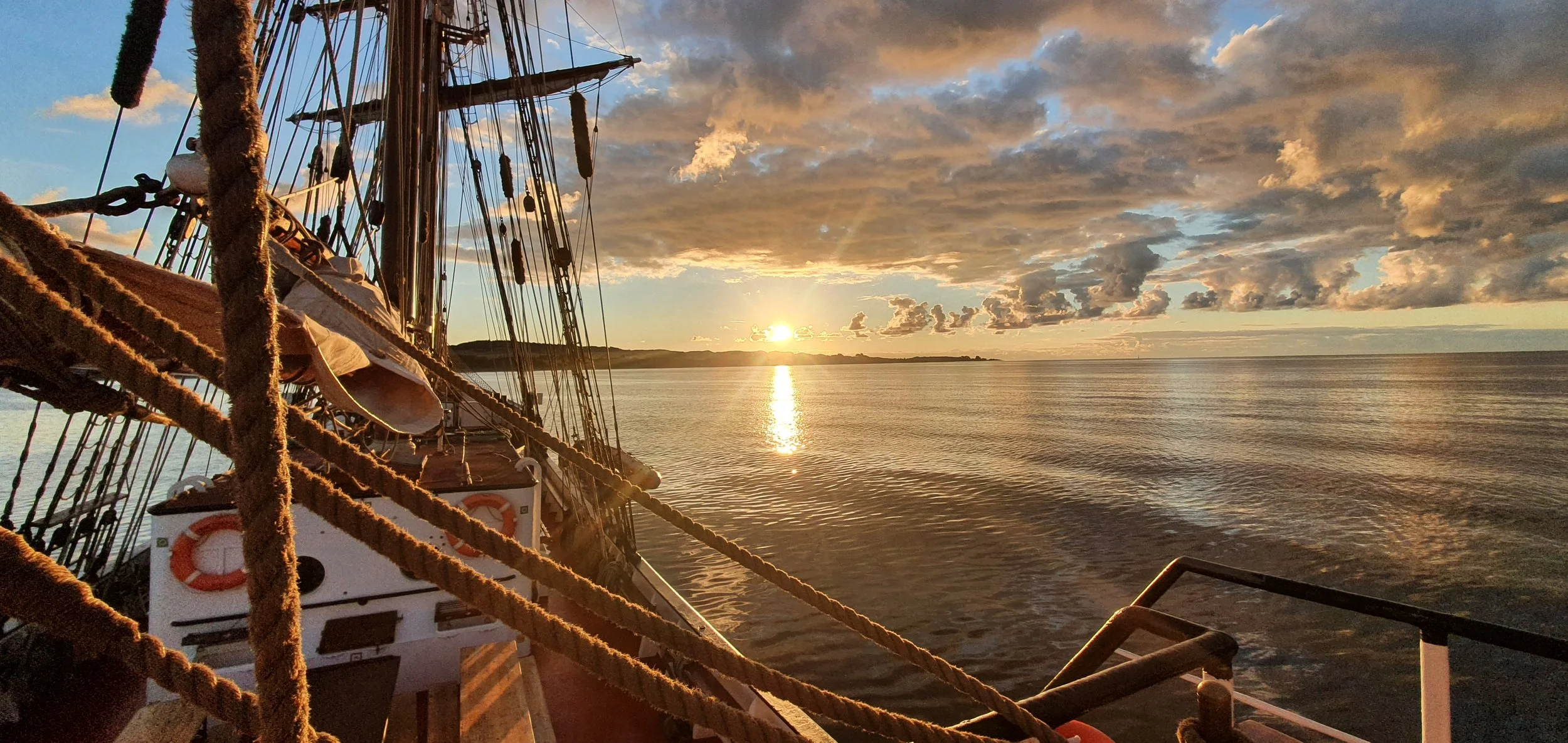 Ein Segelschiff auf ruhigem Wasser bei Sonnenaufgang, mit wolkigem Himmel im Hintergrund.