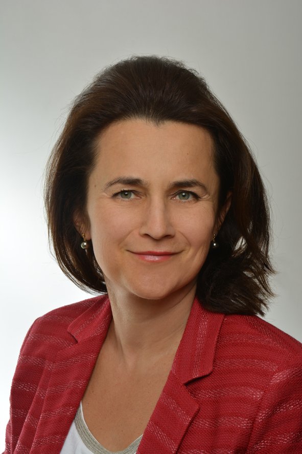 Porträt einer Frau mit braunen Haaren, schwarzem Oberteil und roter Jacke, lächelnd vor neutralem Hintergrund.
