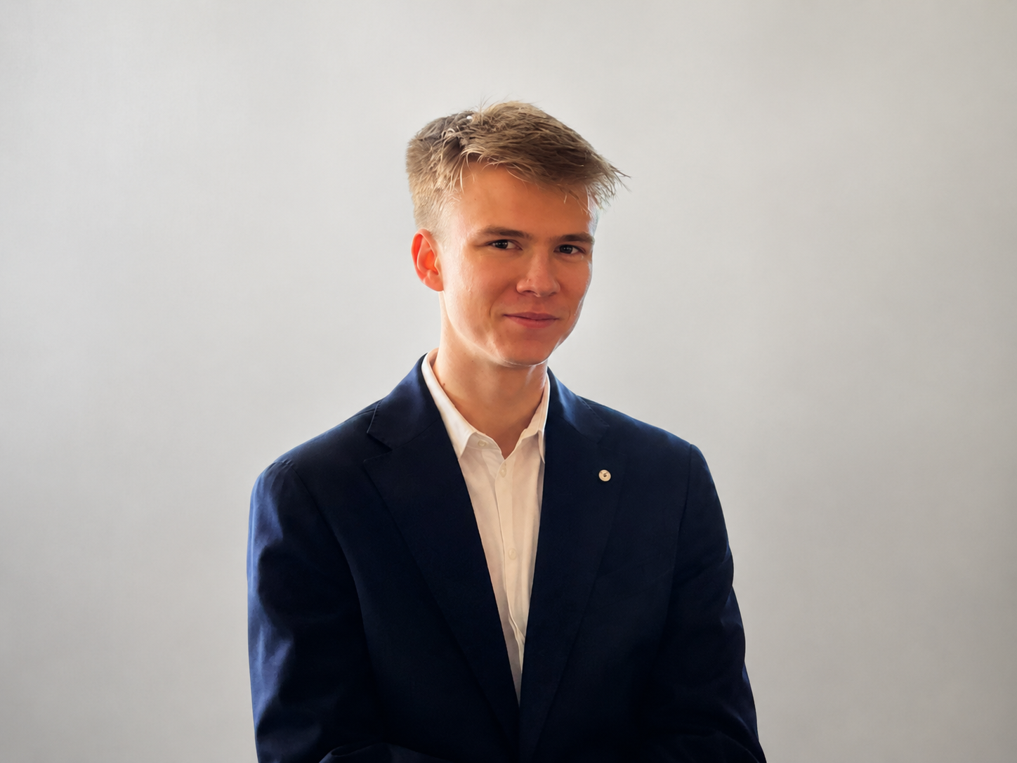 Junger Mann im dunklen Anzug mit weißem Hemd, lächelnd, vor hellem Hintergrund.
