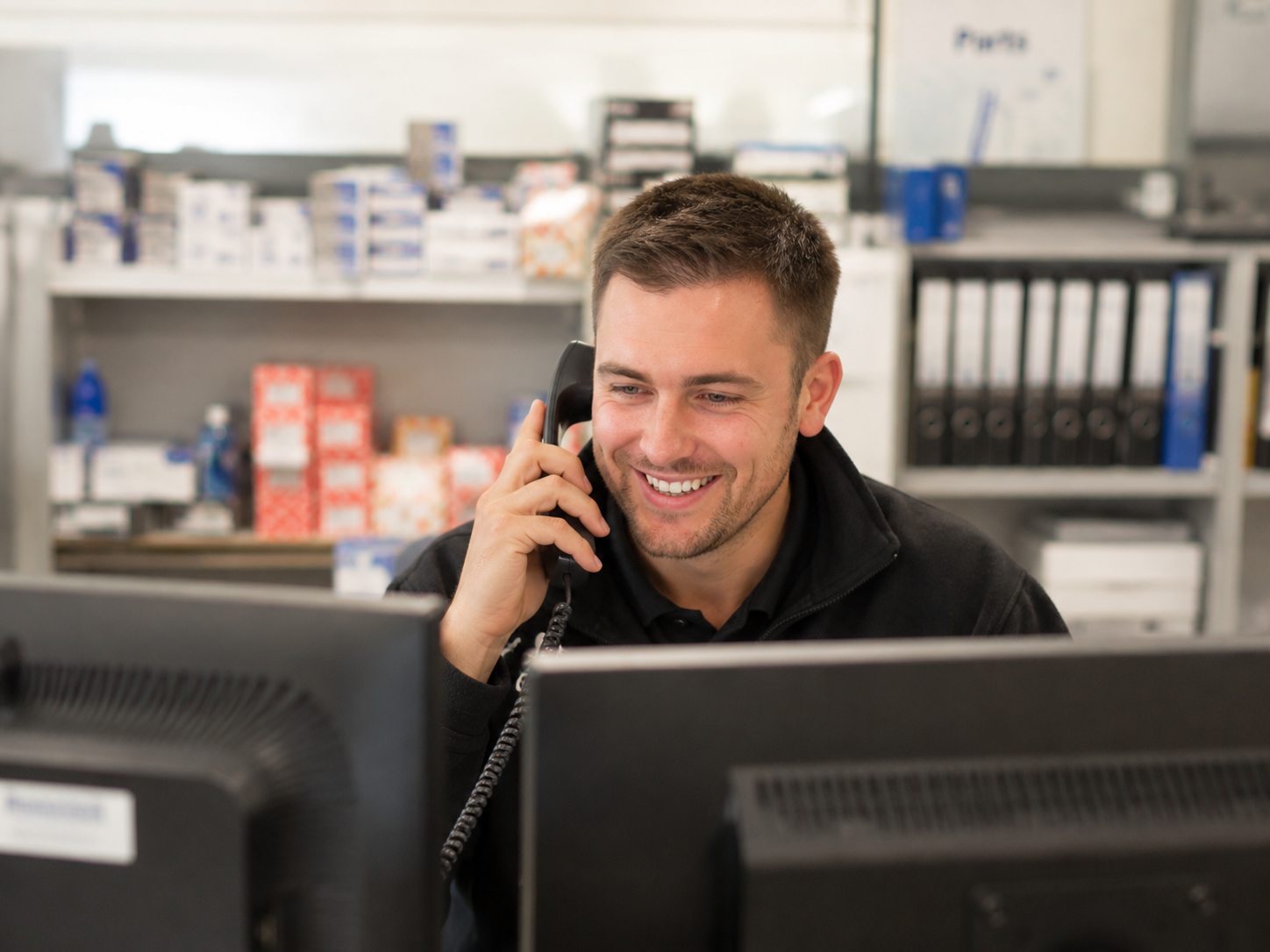 A man smiling while talking on the phone at his desk in front of two computer monitors in an office setting.