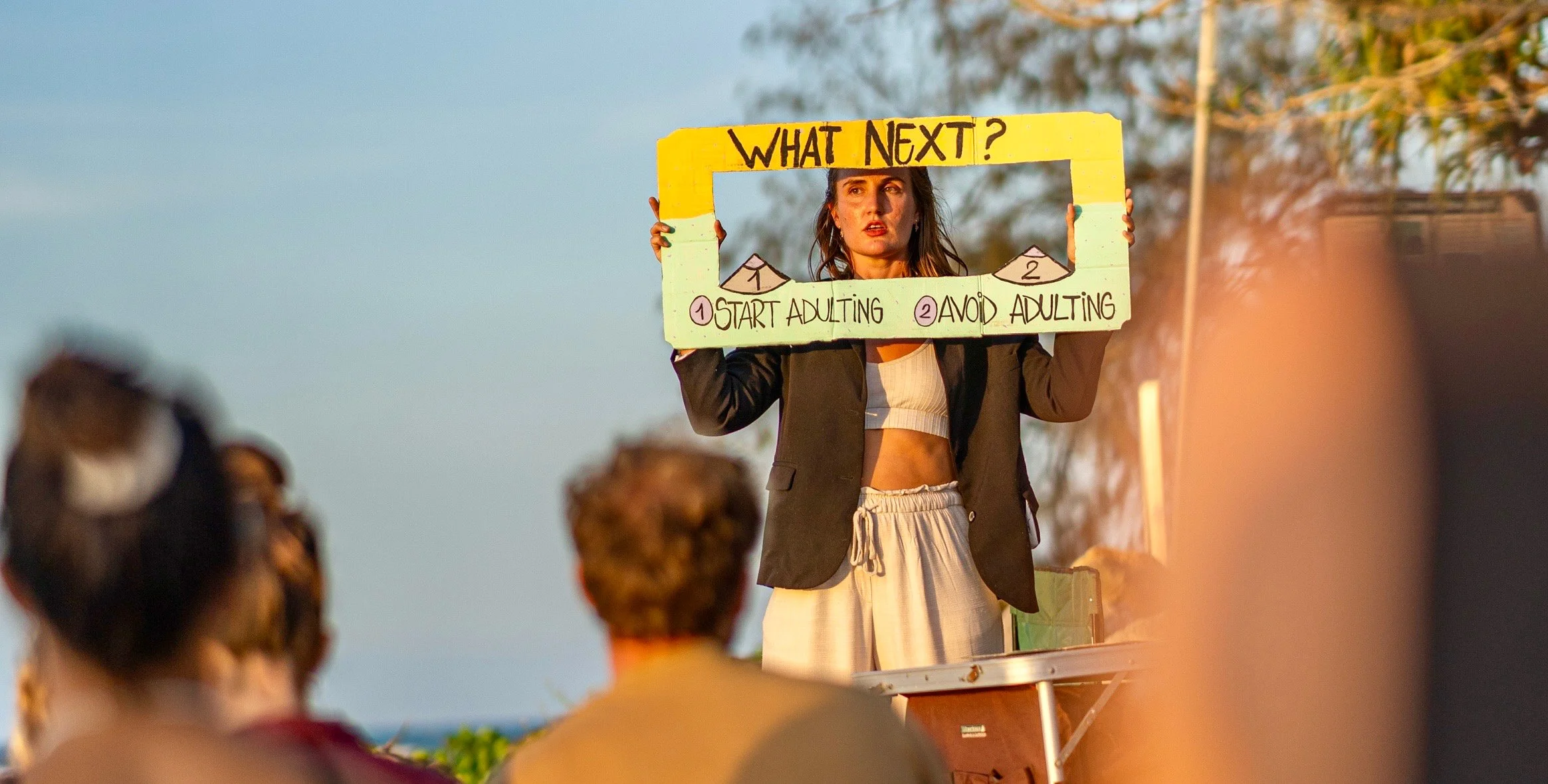 A woman holding a yellow speech bubble-shaped sign that reads 'WHAT NEXT?' with options '1. START ADULTING' and '2. AVOID ADULTING' in front of a crowd outdoors during sunset.