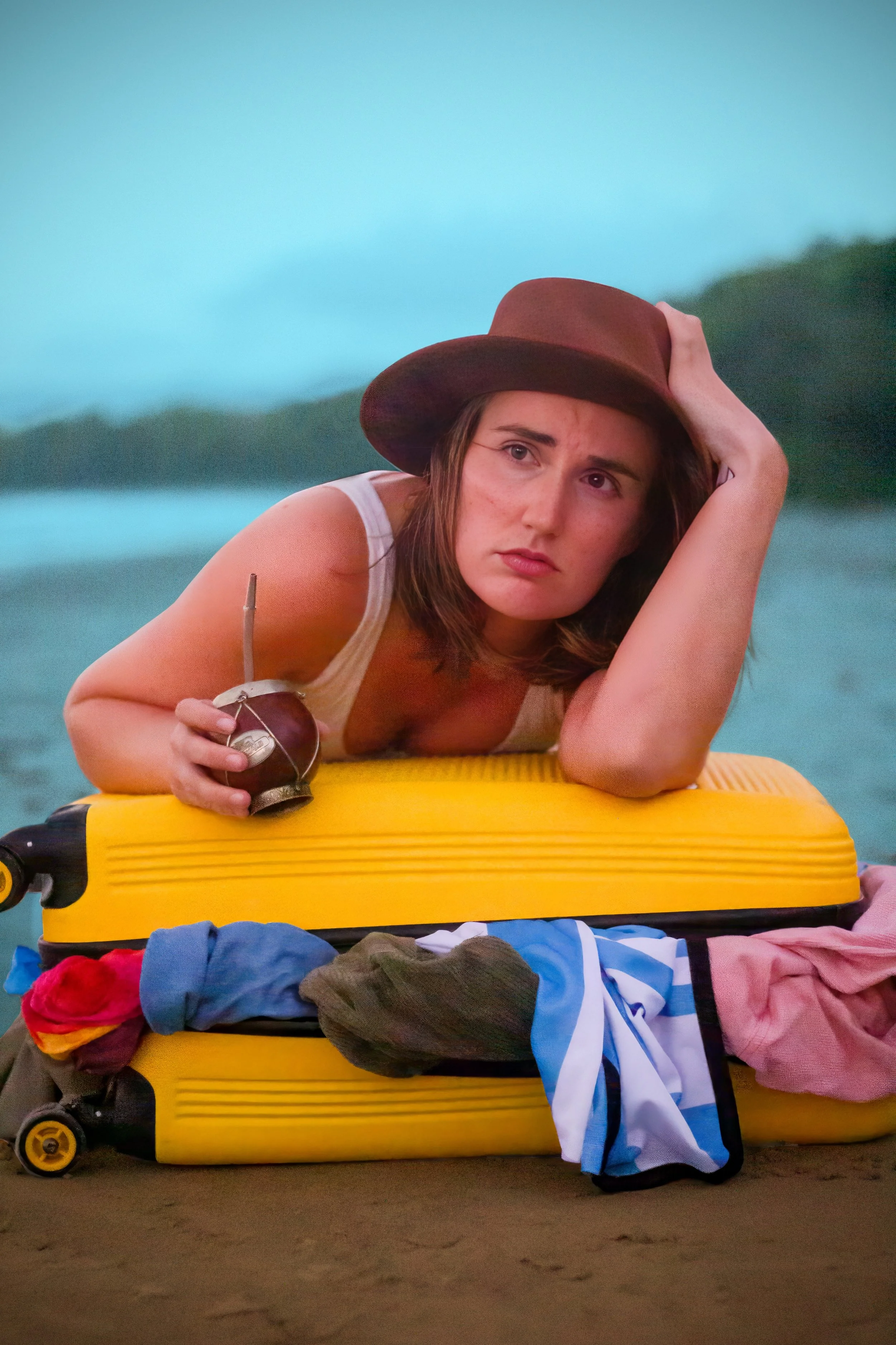 Young woman lying on a yellow suitcase on a sandy beach, wearing a wide-brimmed hat and holding a coconut with a straw. Clothes are scattered around her on the sand, and a body of water and distant trees are in the background.