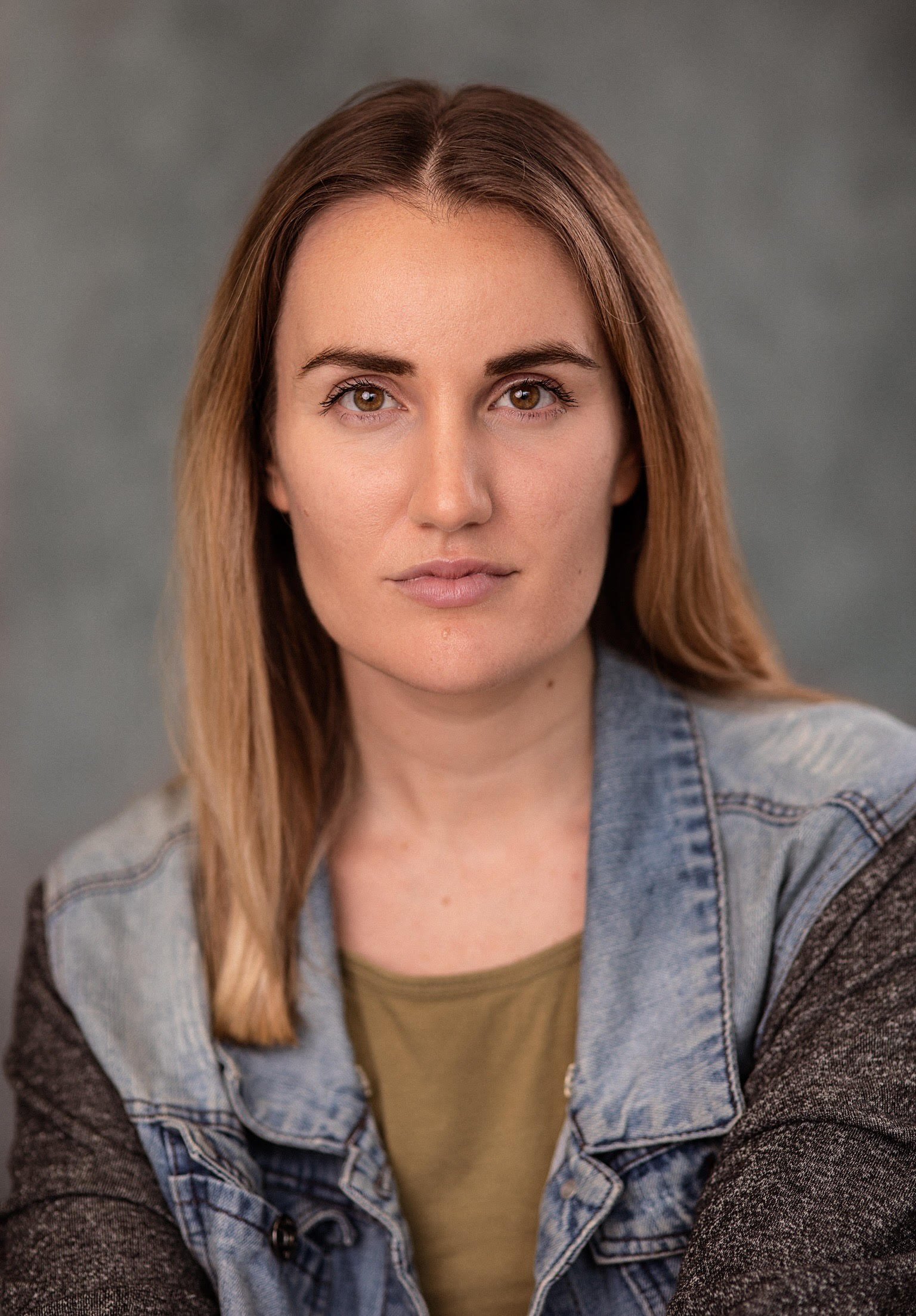 Close-up of a young woman with long, light brown hair, wearing a denim jacket over a beige top, with a neutral background.