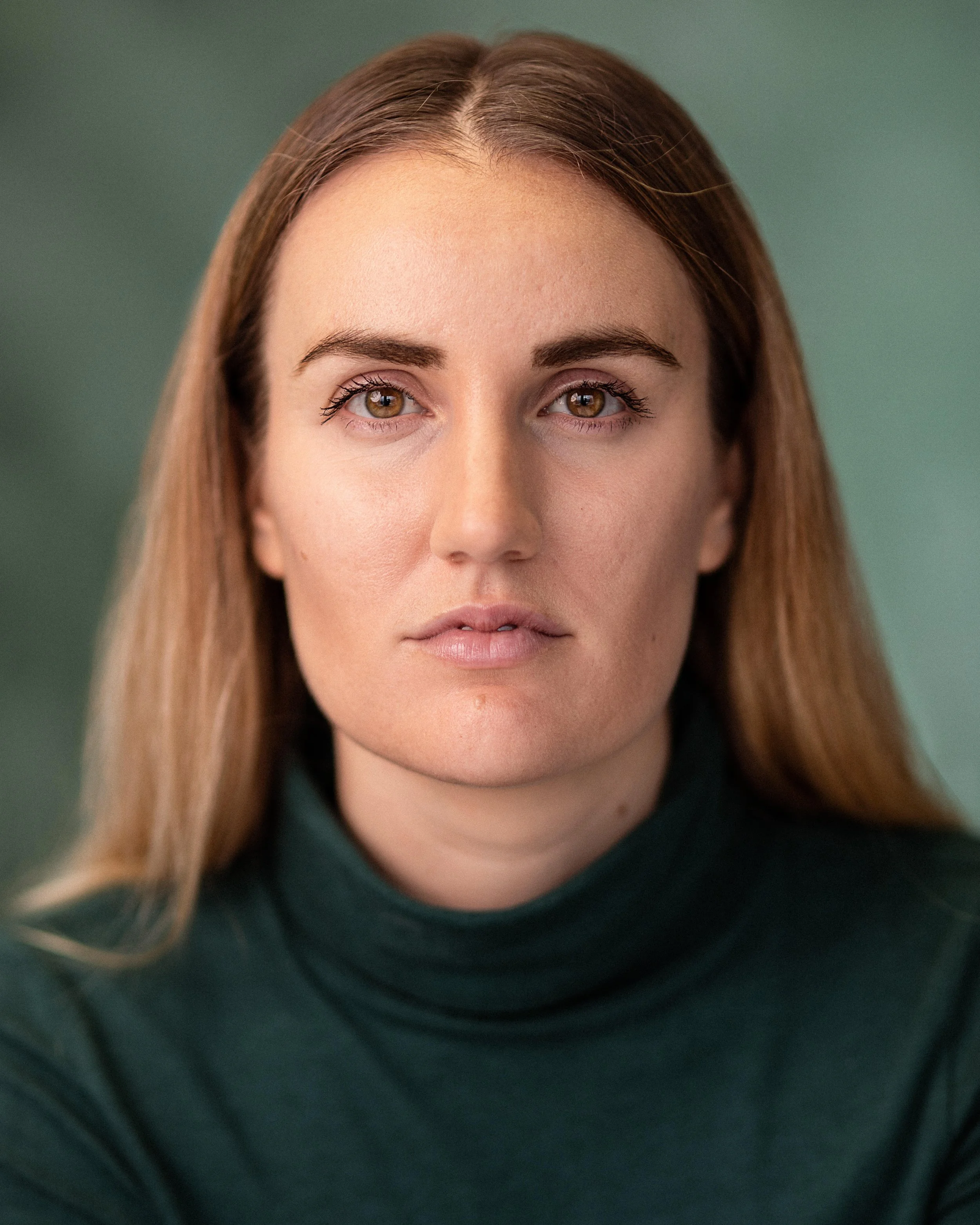 Close-up of a woman's face with long brown hair, brown eyes, and natural makeup, wearing a dark top, against a green background.