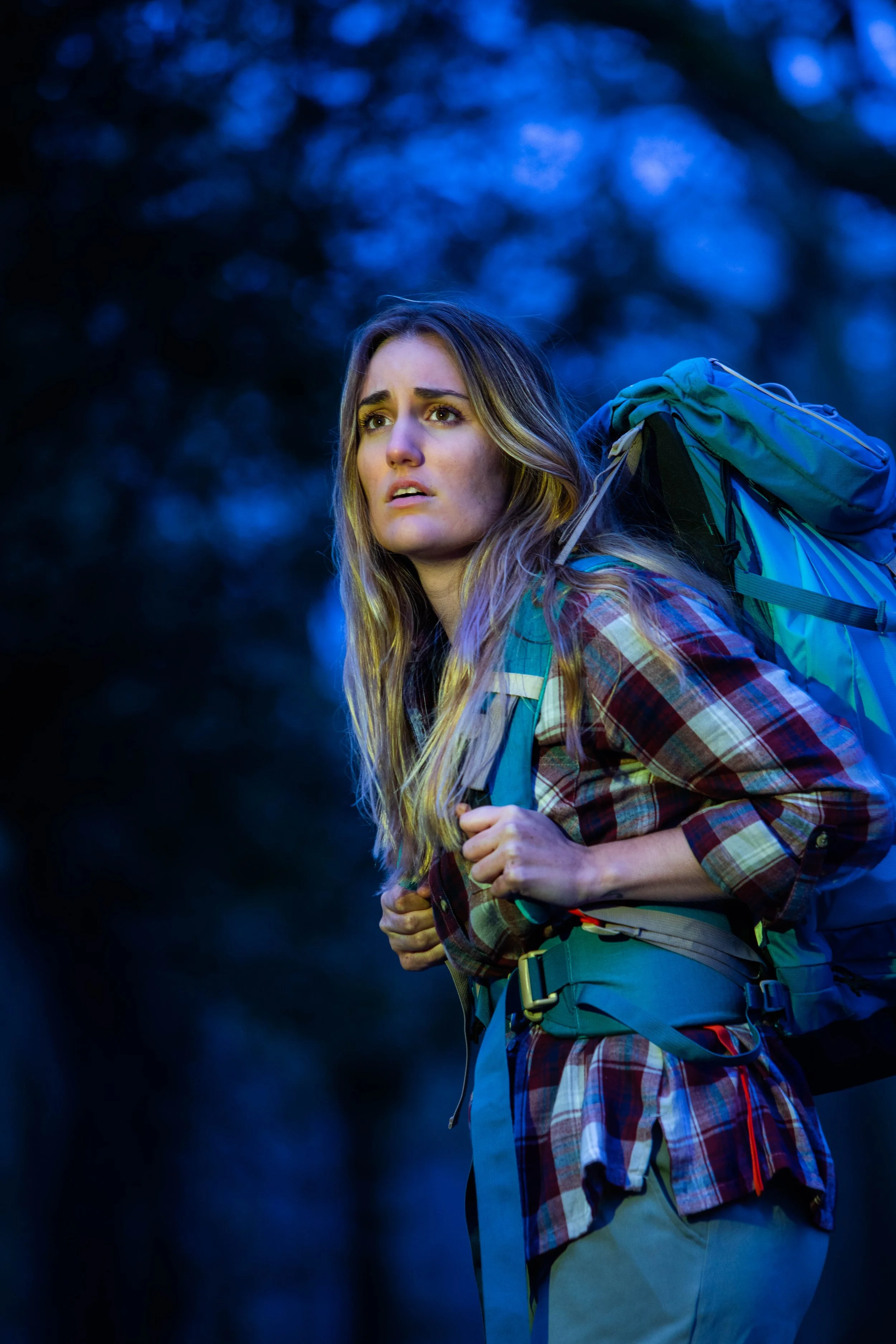 A young woman with long blonde hair wearing a plaid shirt and hiking gear, carrying a backpack, standing outdoors with a dark, blurred forest background during evening or dawn.