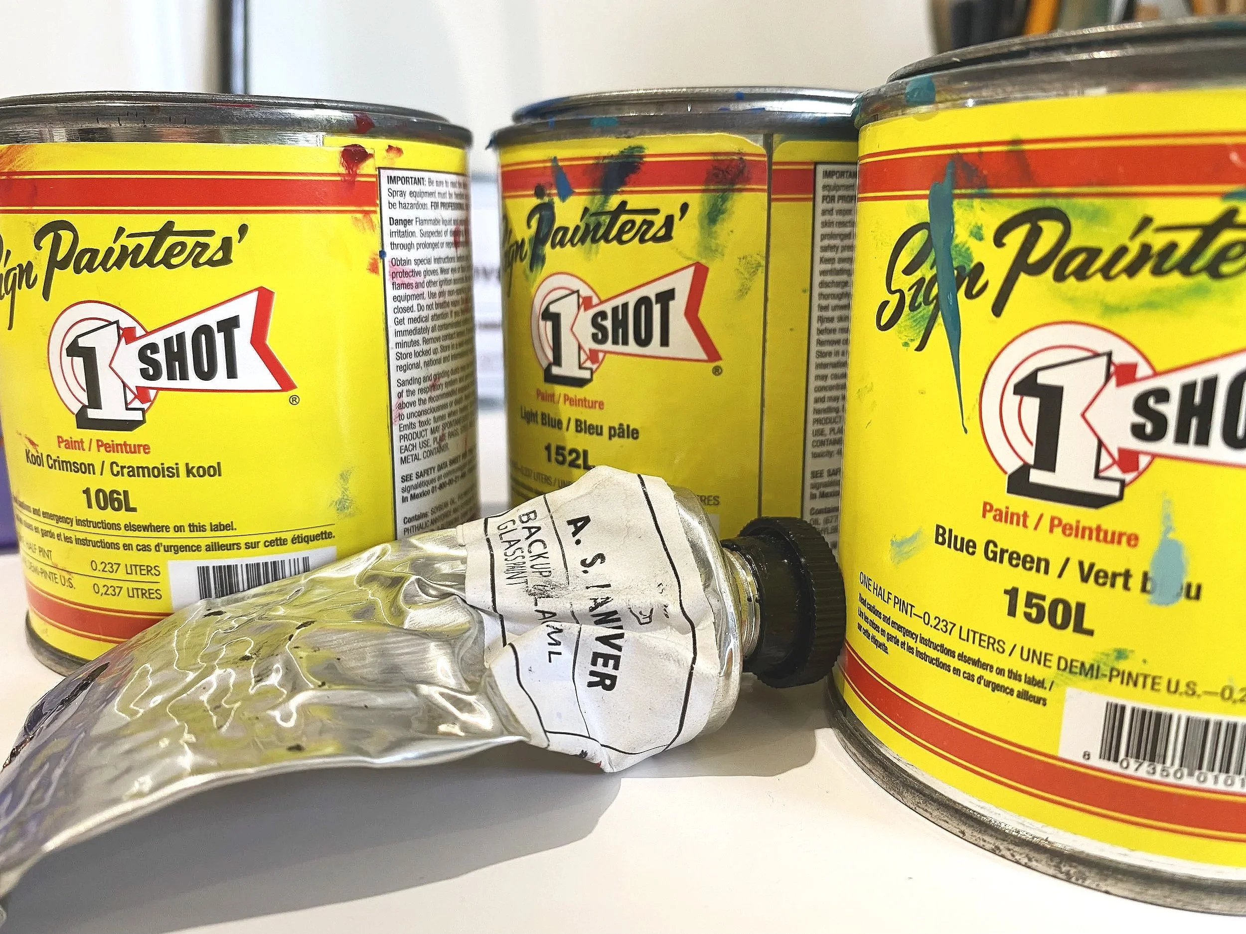 Close-up of three cans of sort paint labeled '1 Shot' with a yellow background and colorful splashes, and a crumpled plastic bottle and a small tube in front.