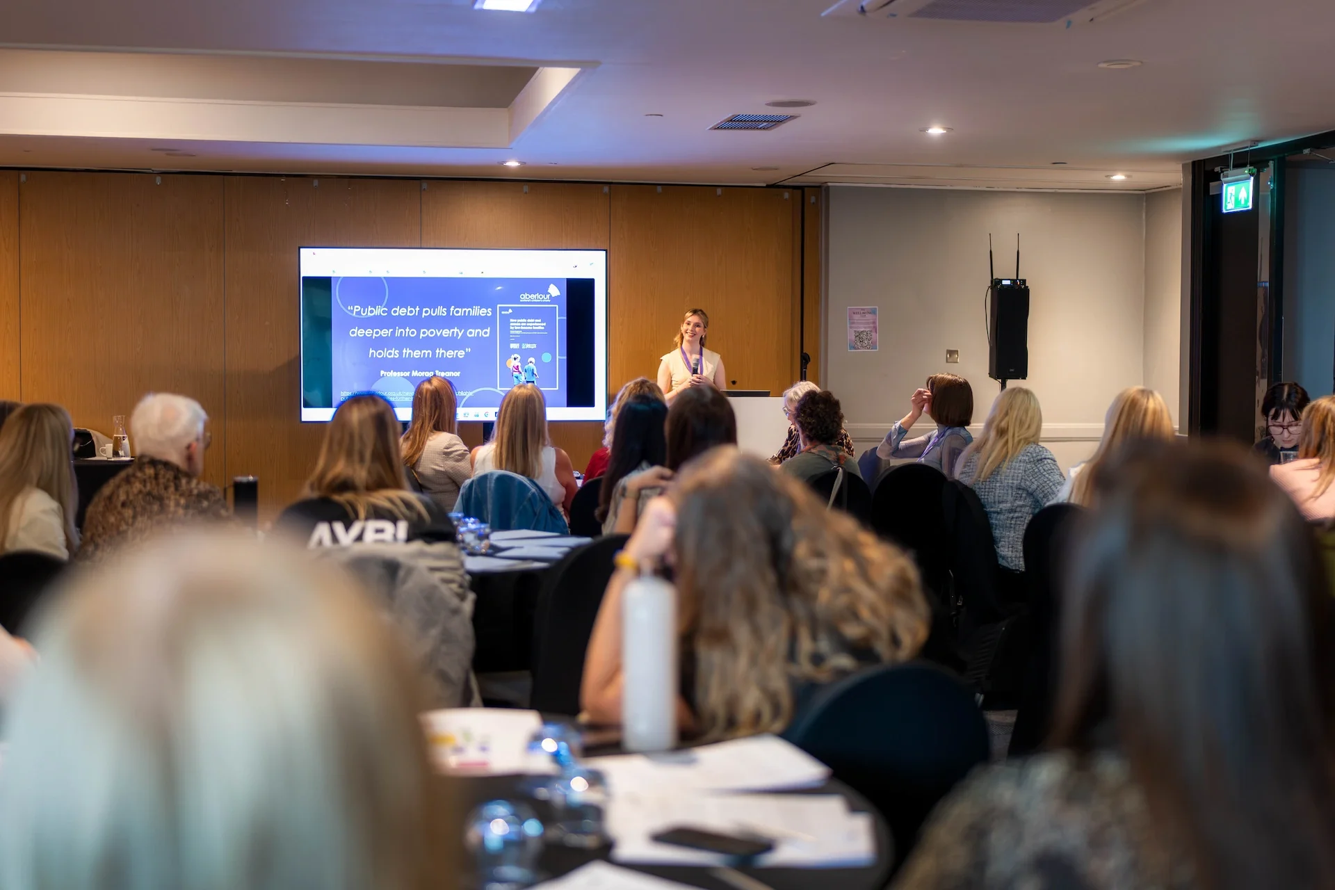 A woman presenting at a conference in front of a large screen with a quote about public debt, surrounded by seated audience members in a conference room.