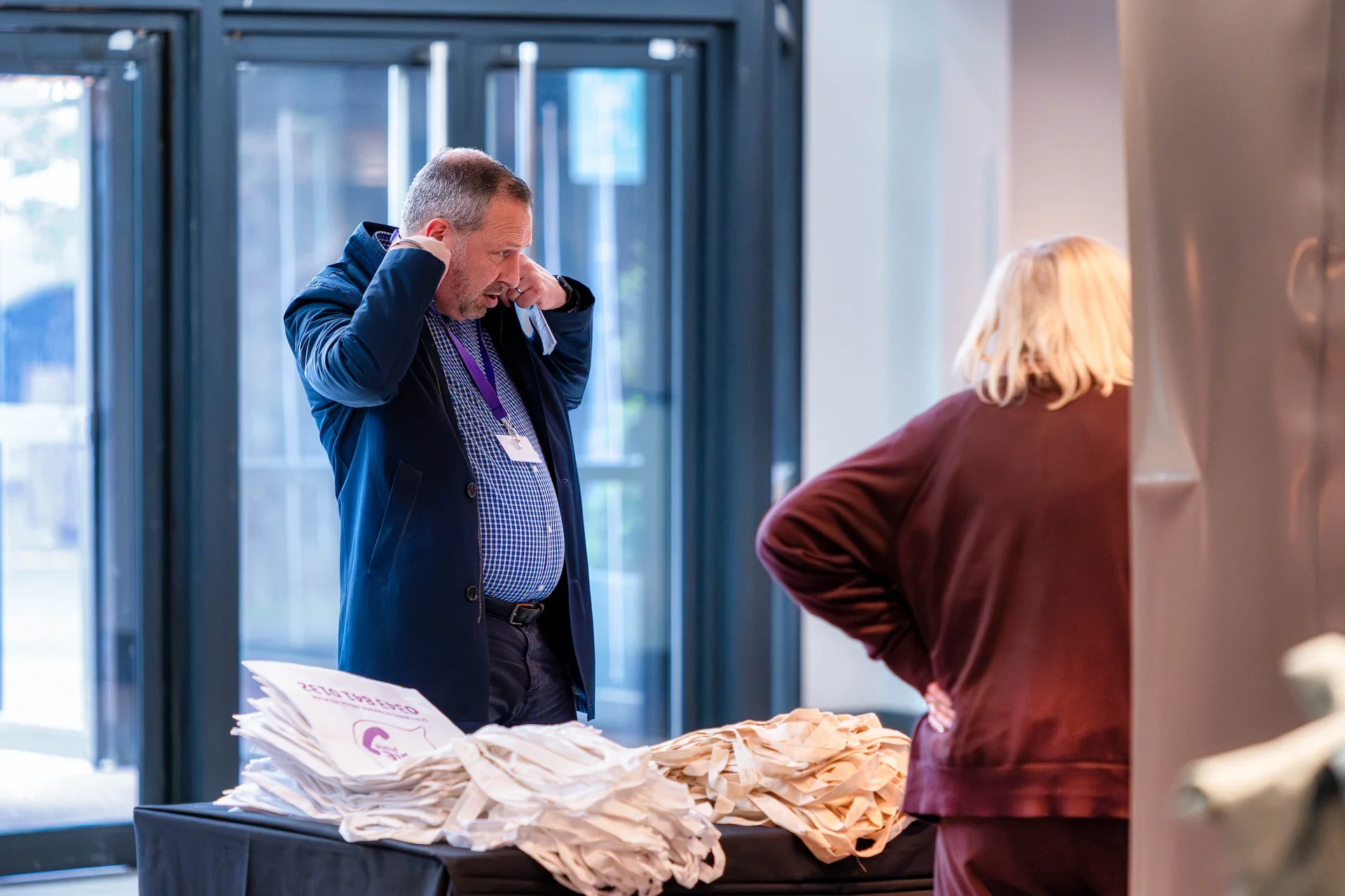 A man with short hair and a beard, wearing a blue blazer and checkered shirt, is adjusting his hearing aids while talking to a woman with blonde hair, wearing a brown jacket, at a registration table filled with paper bags.