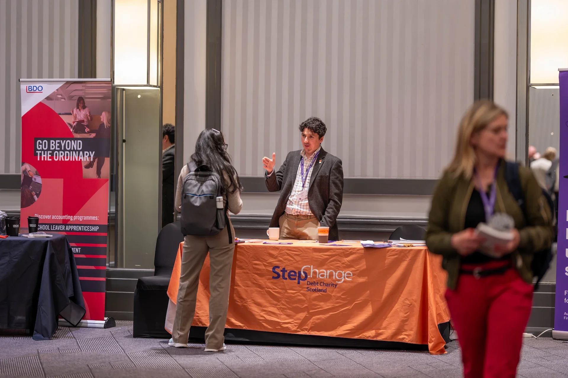 A group of people at a conference or event, with one man talking to a woman with a backpack at a booth. The table has a bright orange tablecloth with the logo 'Step Change' and the text 'Debt Charity Scotland.' There are banners on either side of the booth, one reading 'Go Beyond the Ordinary' and the other with promotional information. The background features a conference room with vertical striped walls.