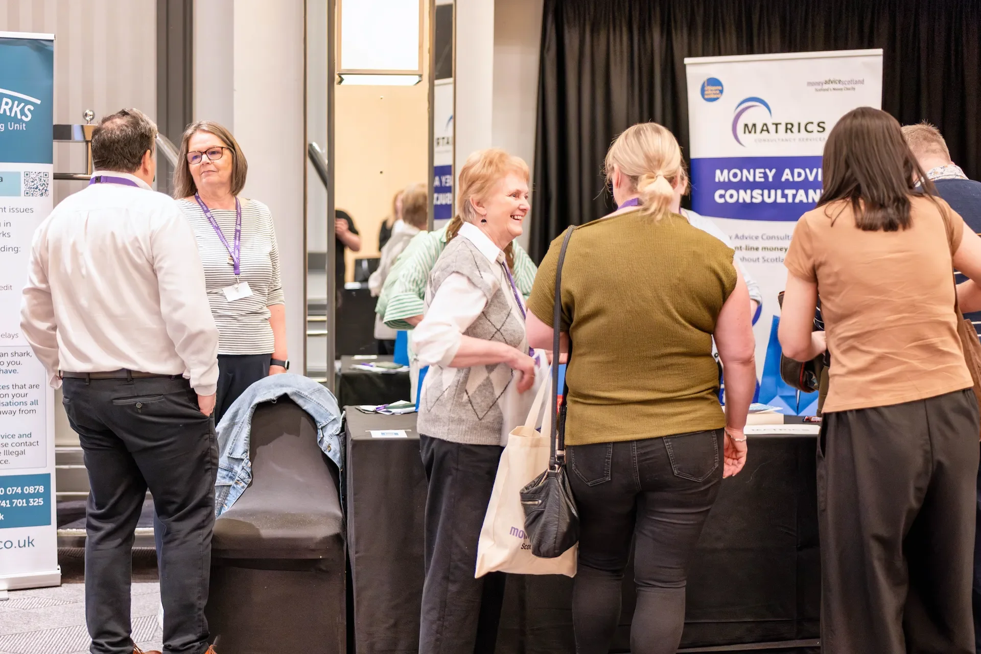 People attending a financial advice conference, engaging in conversations around a booth that displays banners for money advisory services.
