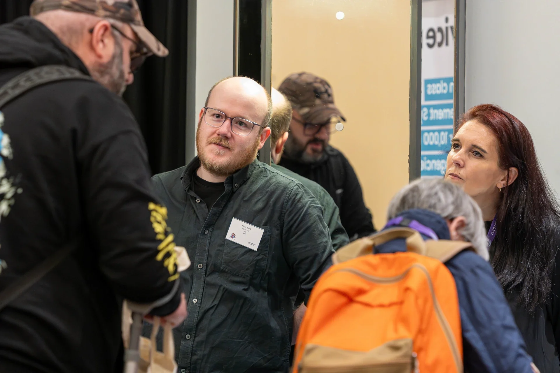 People engaging in conversation at a professional event or conference, with a focus on a man wearing glasses and a name tag, and a woman with long dark hair, in an indoor setting.