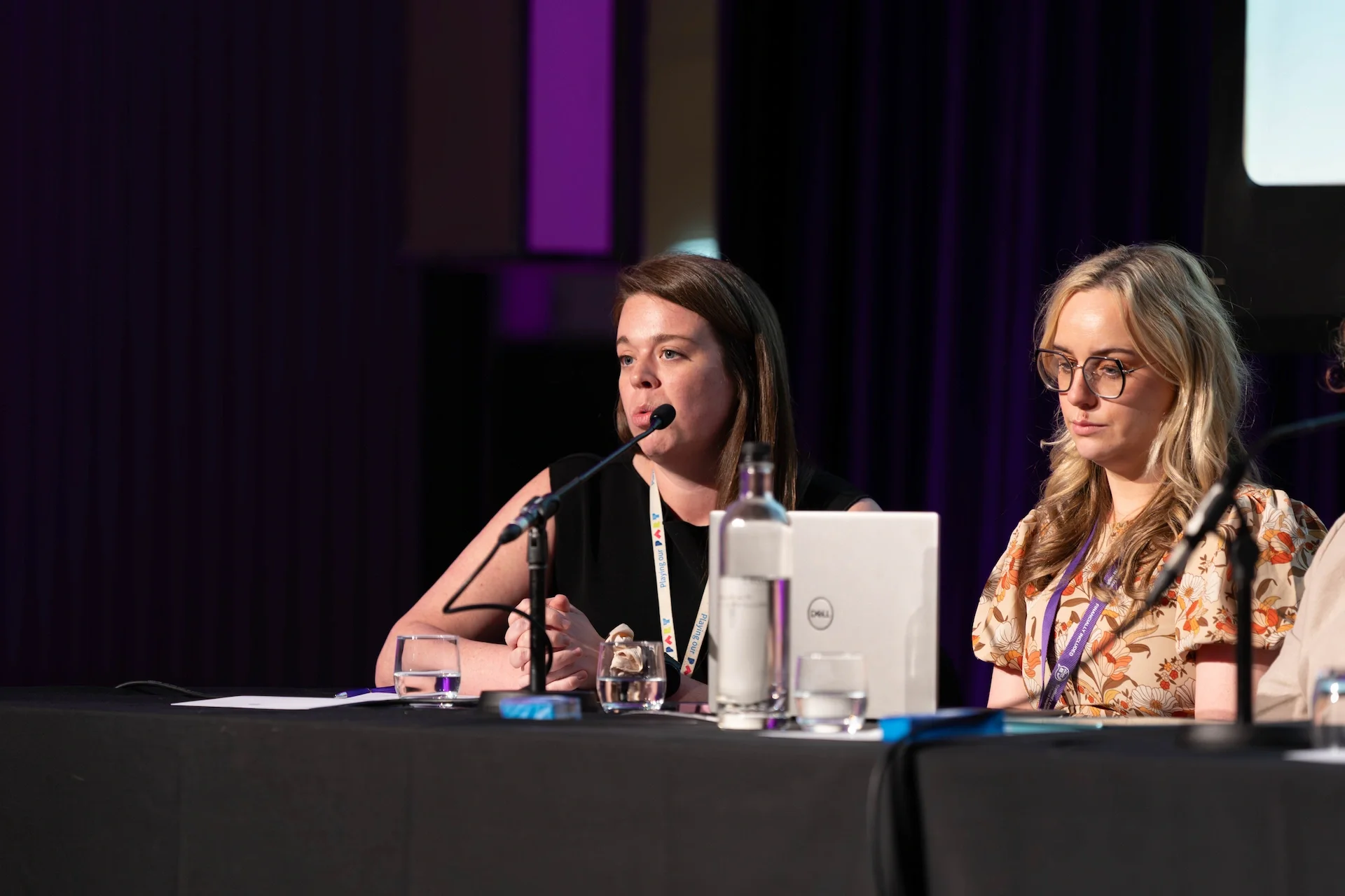 Two women seated at a table during a conference or panel discussion, with one woman speaking into a microphone and the other listening, surrounded by glasses of water and electronic devices.