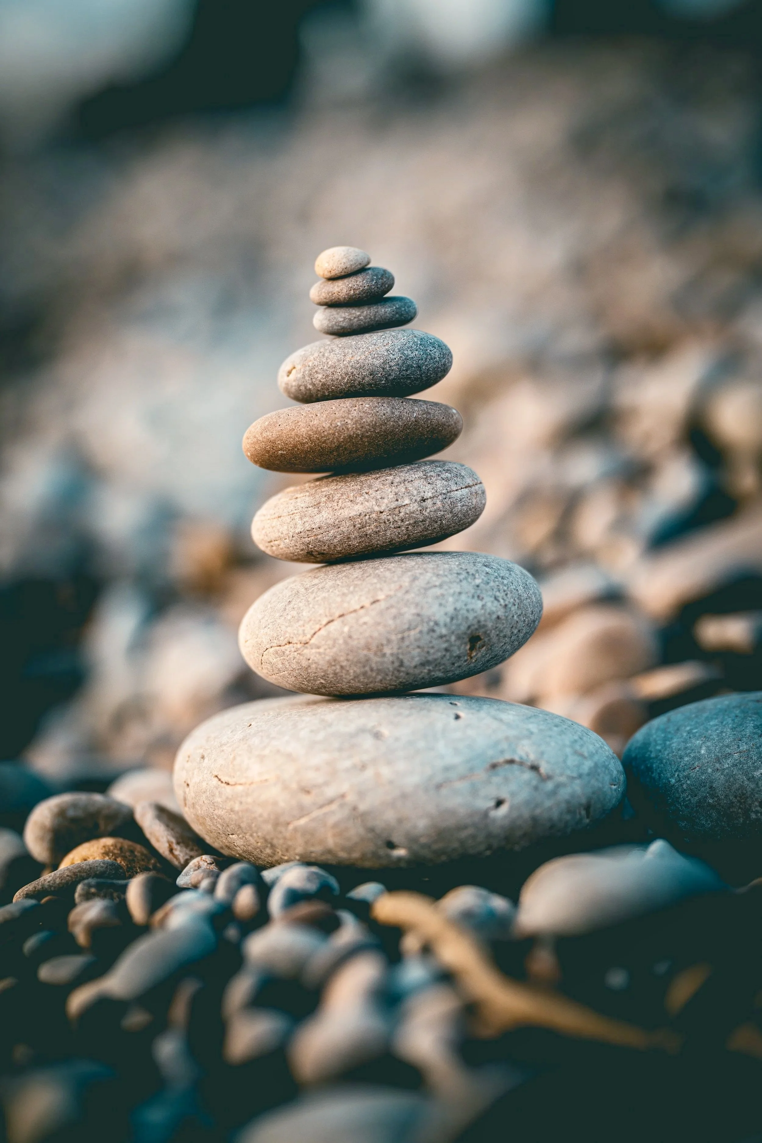 A stack of seven smooth, flat stones balanced on top of each other in a natural outdoor setting with a blurred background of smaller rocks and pebbles.