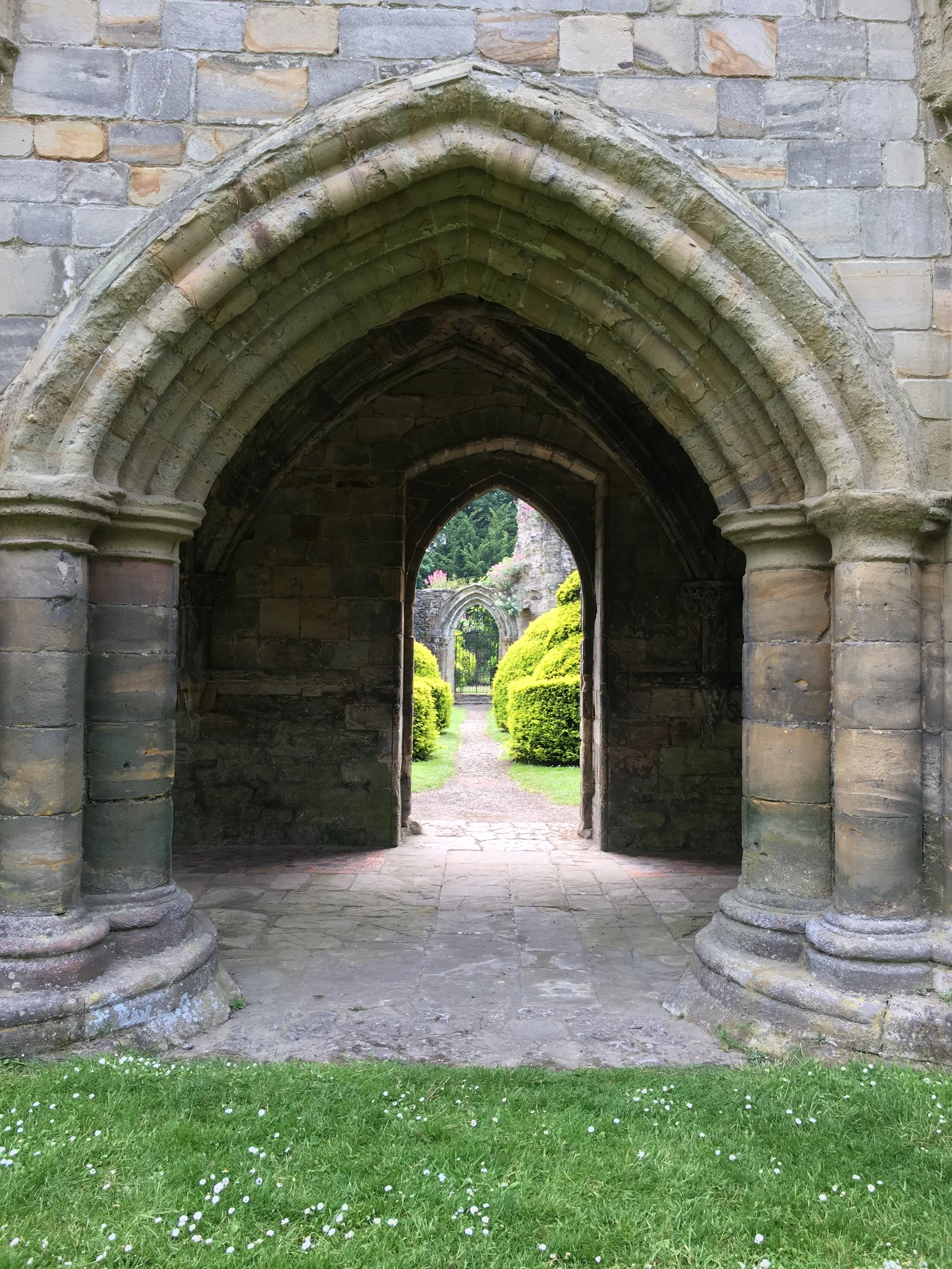 Stone archway view with green grass and white flowers in foreground, leading to a garden with bushes and flowered trees