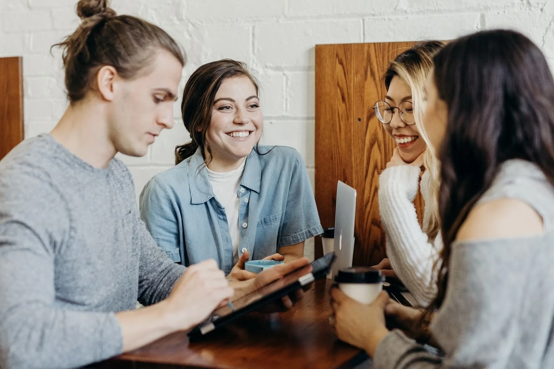 Four young women sitting at a wooden table, smiling, with a white brick wall behind them, engaged in conversation and using electronic devices.