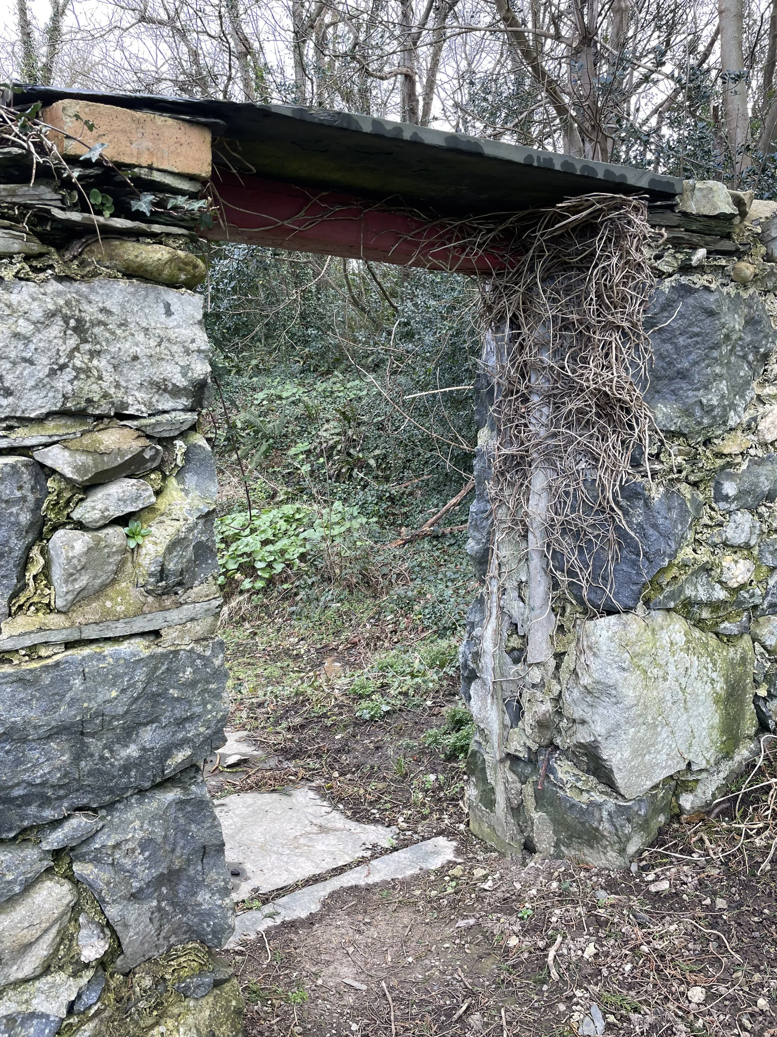 A stone and brick archway with a wooden beam overhead, situated in a wooded area with trees and plants in the background.