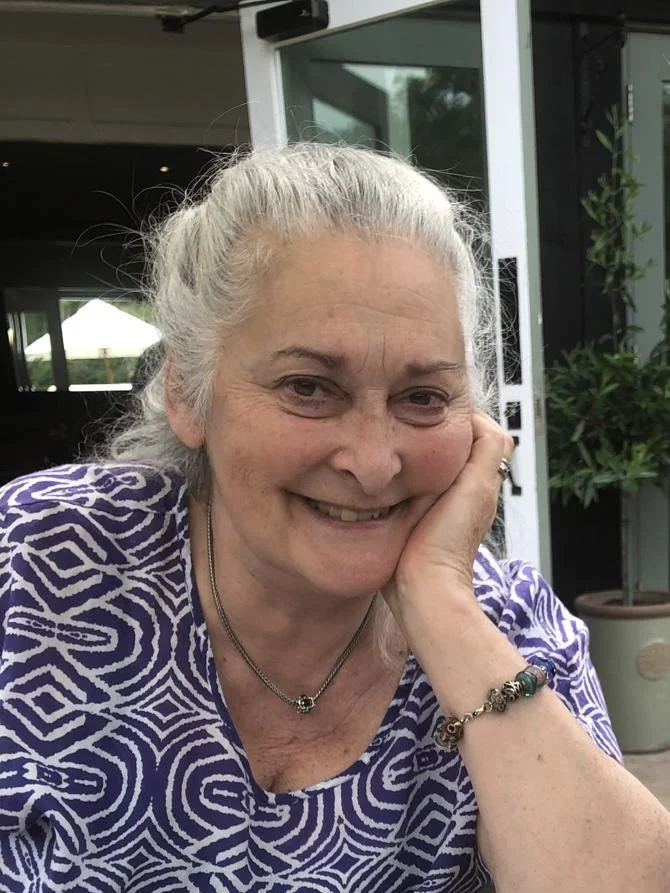 A smiling elderly woman with gray hair tied back, wearing a patterned purple and white top, a necklace, and a bracelet, resting her face on her hand in an outdoor setting.