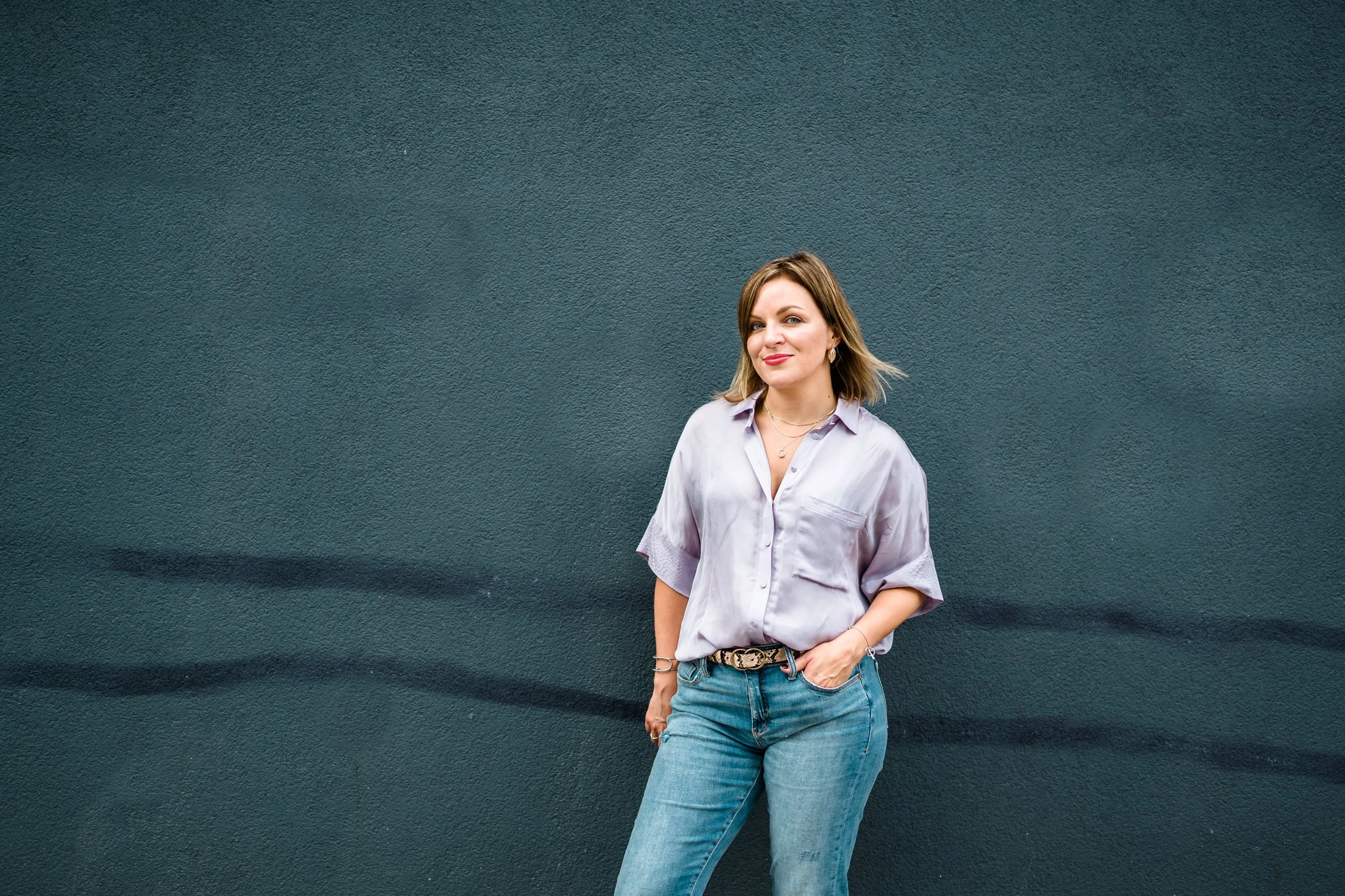 A woman with shoulder-length hair and light skin, smiling softly, standing against a plain dark blue wall. She is wearing a light-colored button-up shirt, blue jeans, and some jewelry, with her right hand in her pocket.