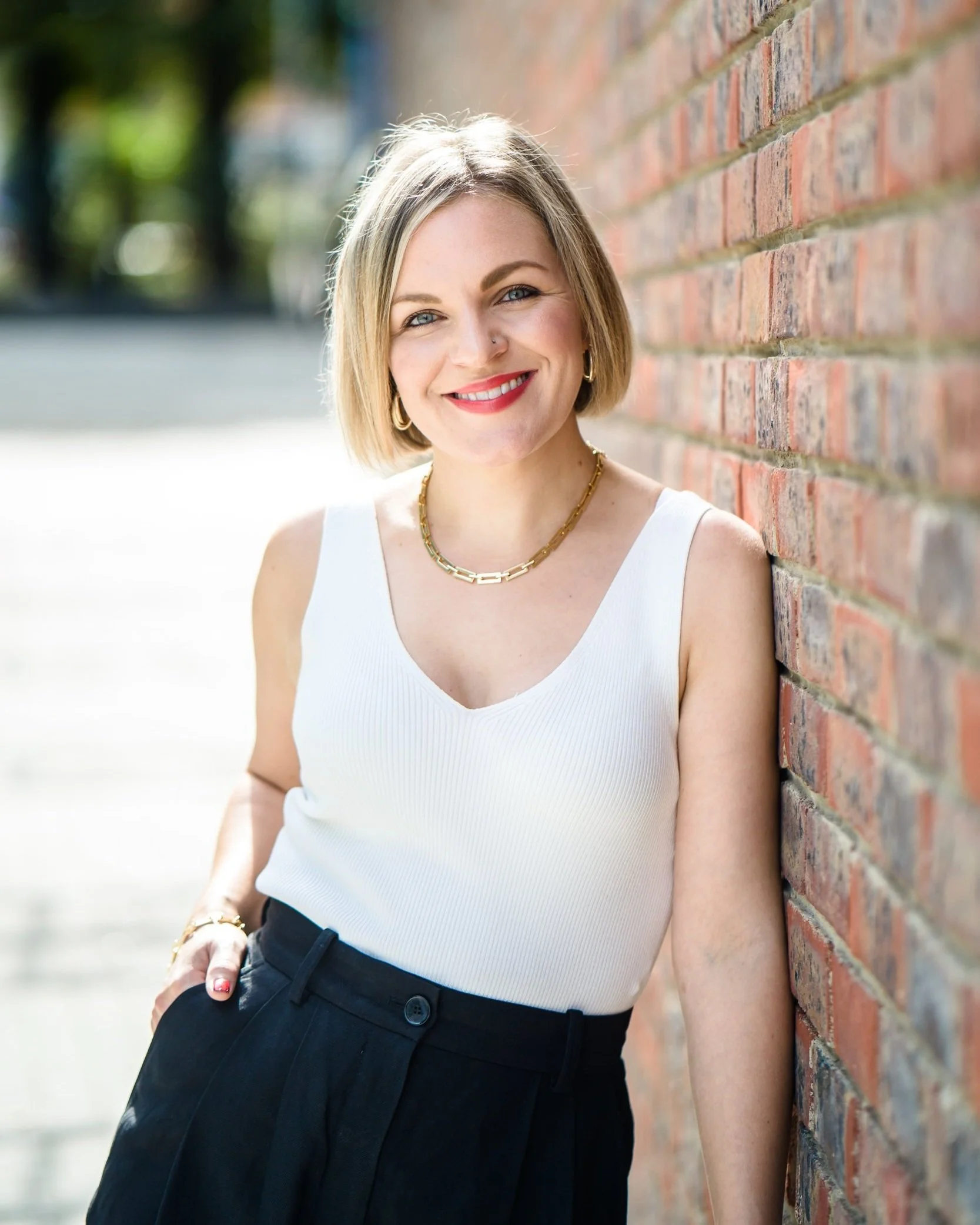 A woman with shoulder-length blonde hair leaning against a brick wall outdoors, smiling at the camera. She is wearing a white sleeveless top, black high-waisted pants, gold jewelry including a chain necklace and hoop earrings, and has a nose ring.