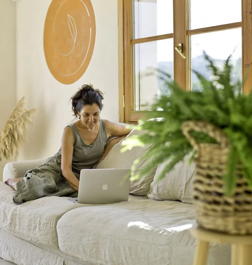 A woman sitting on a cream-colored sofa in a bright room, using a laptop, with a window behind her and a potted plant in the foreground.