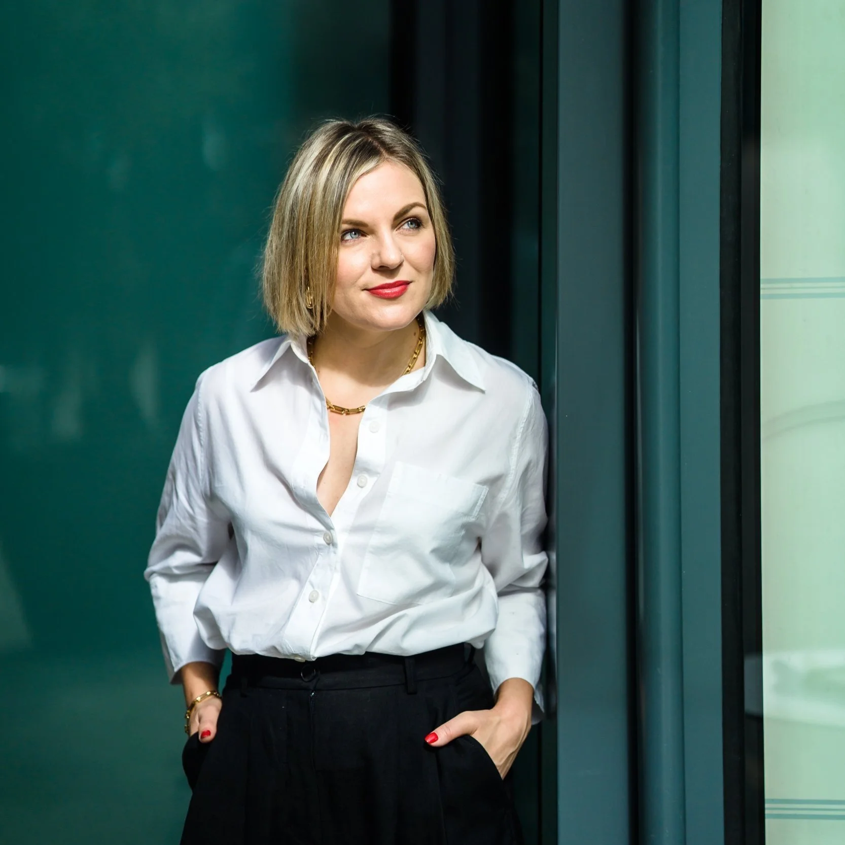 A woman with blonde hair, wearing a white button-up shirt and black pants, standing indoors by a large window with a reflective teal wall in the background. She has red lipstick, gold jewelry, and is looking off to the side with a slight smile.