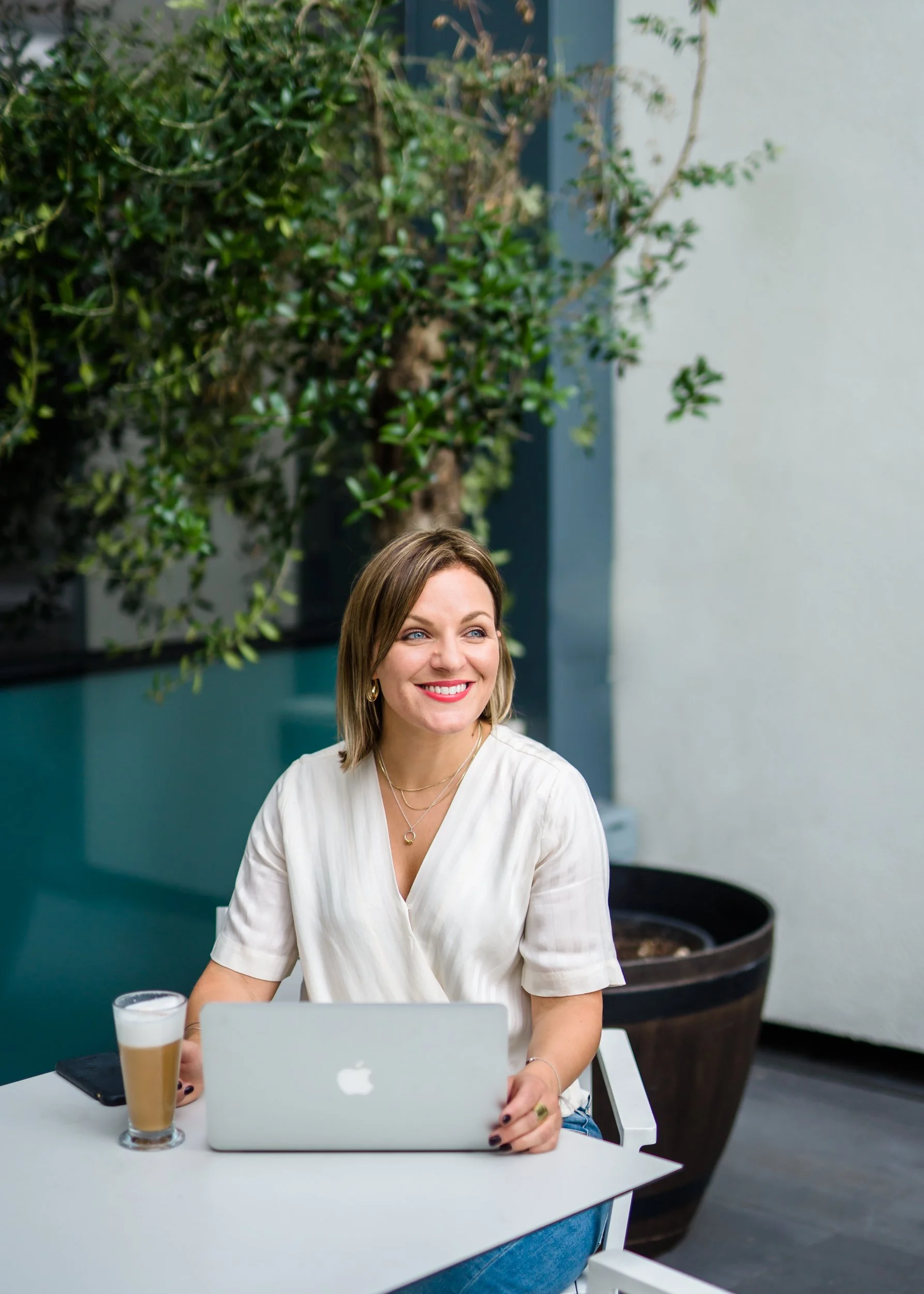 A woman sitting at a white table outdoors, smiling, with a glass of iced coffee, a laptop, and a smartphone in front of her. There are large potted plants and a gray wall in the background.