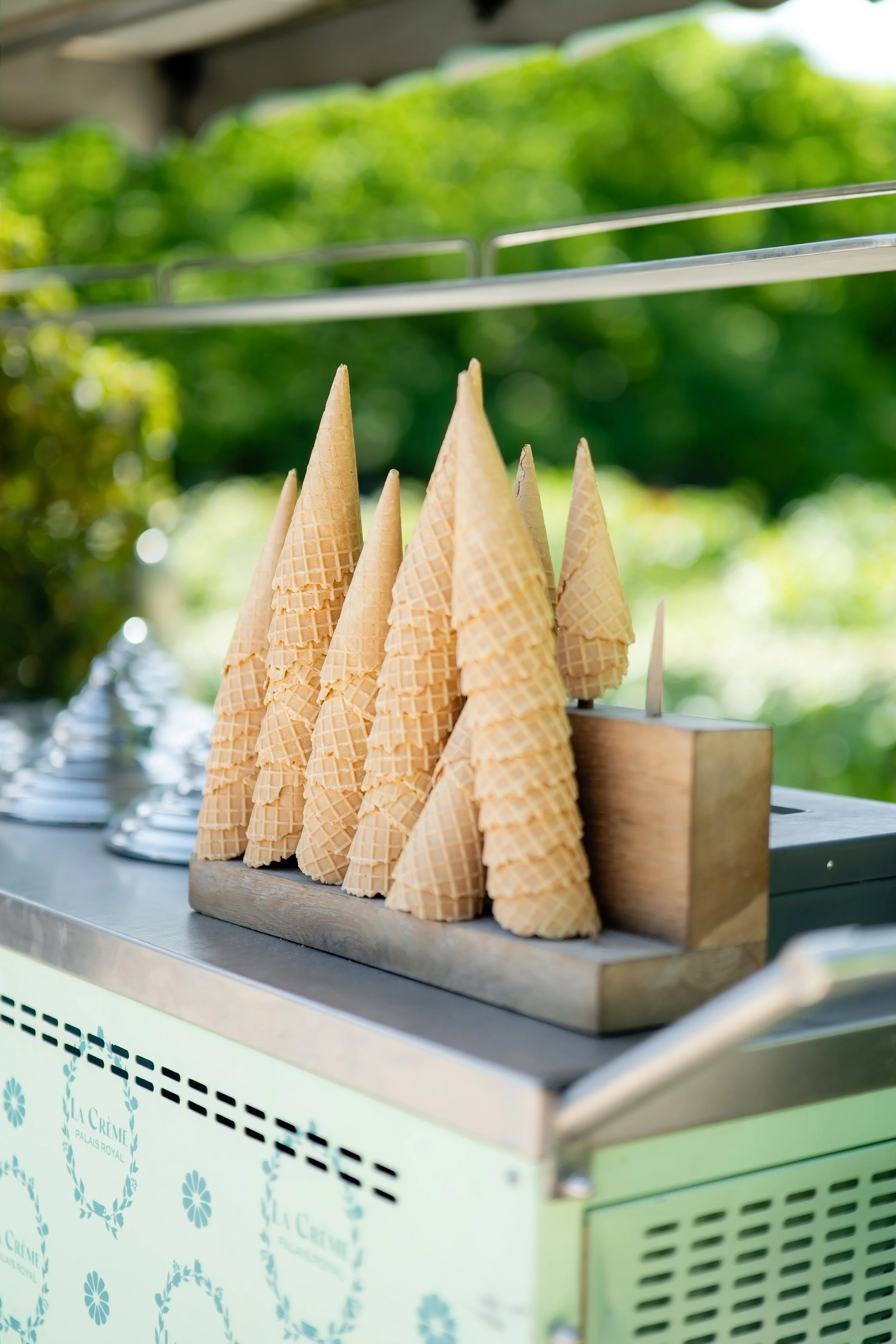 Multiple ice cream cones arranged in a row on a wooden counter, with a blurred green outdoor background.