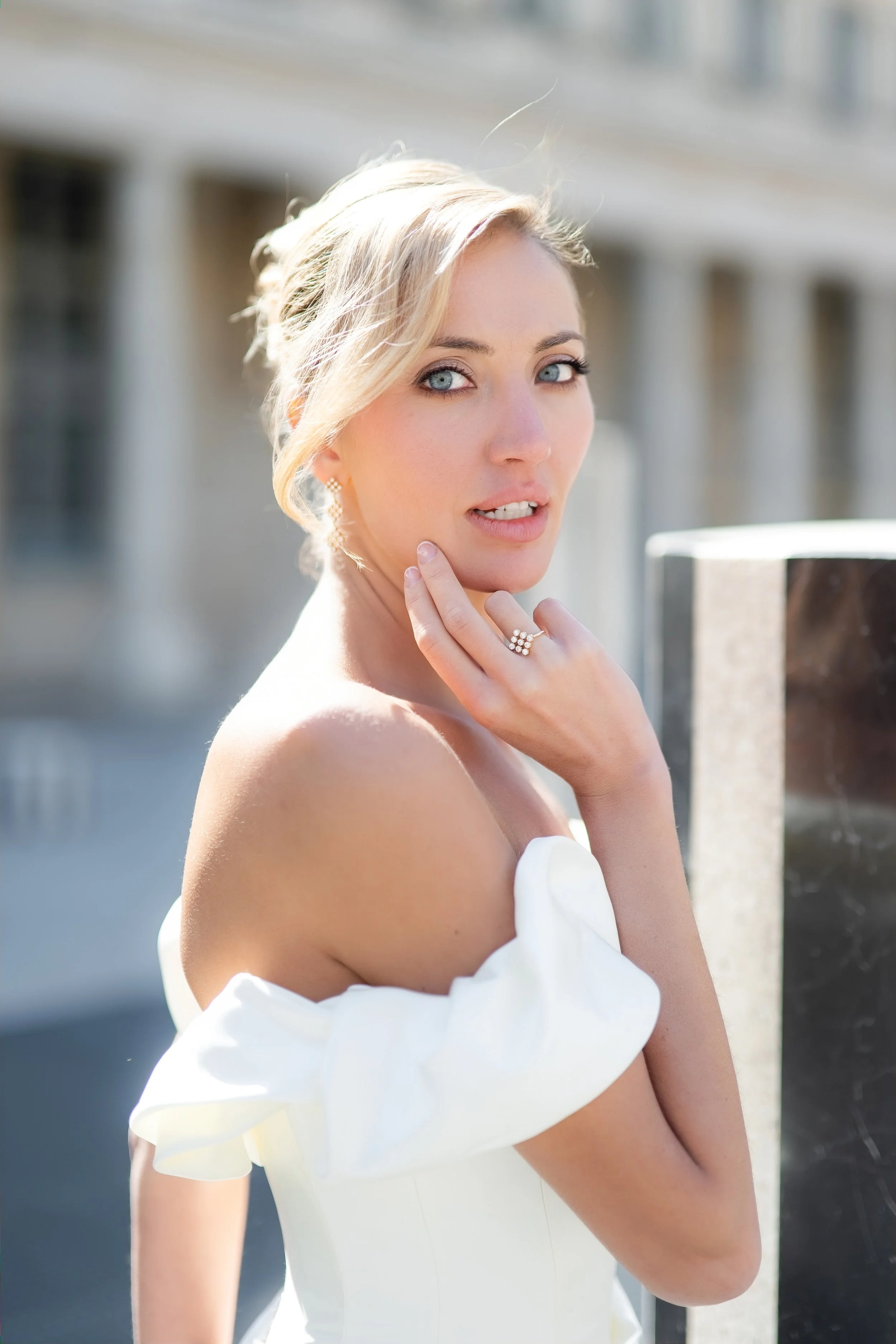 A woman with blonde hair and blue eyes, wearing a white off-shoulder dress and jewelry, posing outdoors on a sunny day.