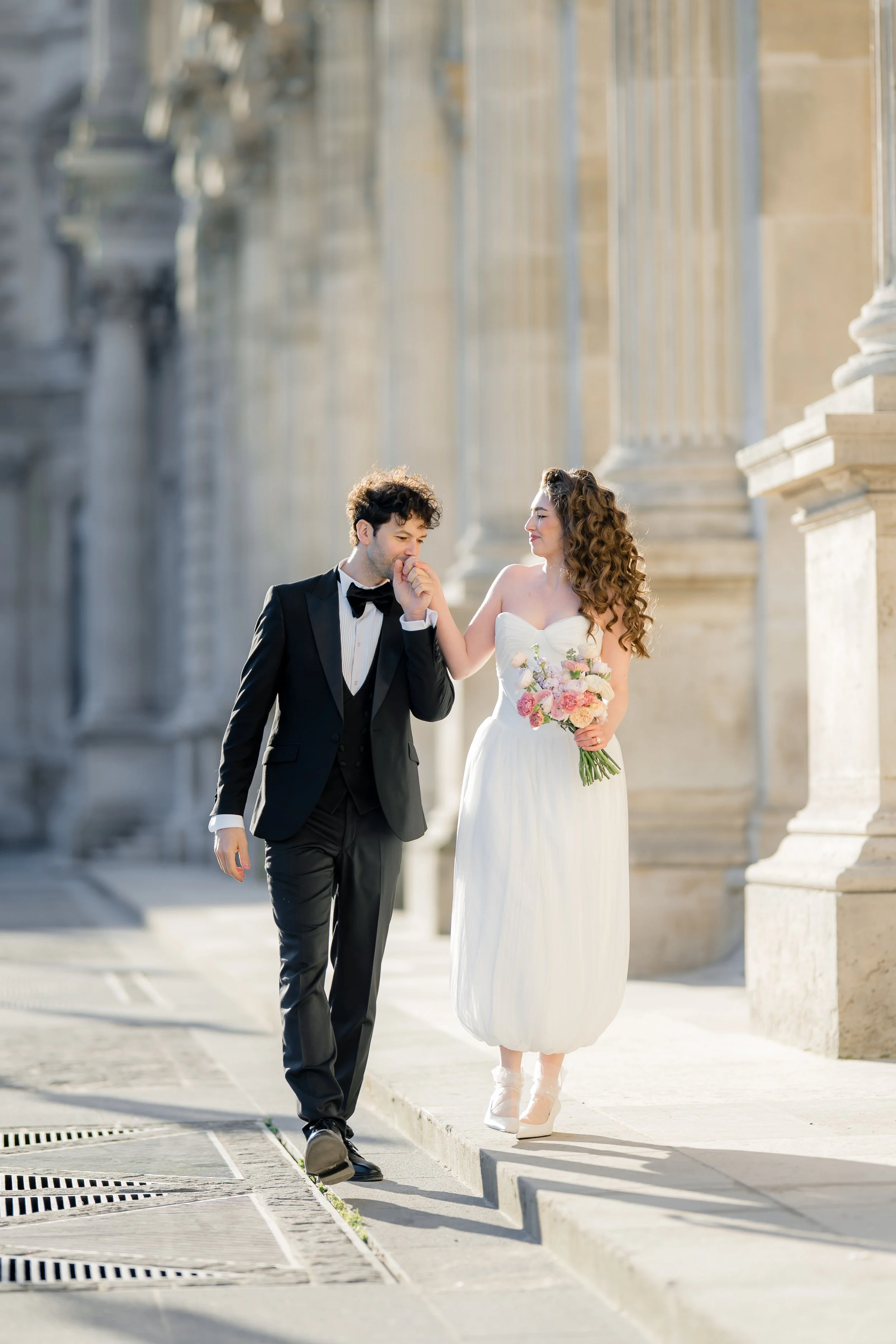 A bride and groom walking together outdoors near a stone building with columns, with the bride holding a bouquet of flowers, and the groom wearing a tuxedo, smiling as they walk.