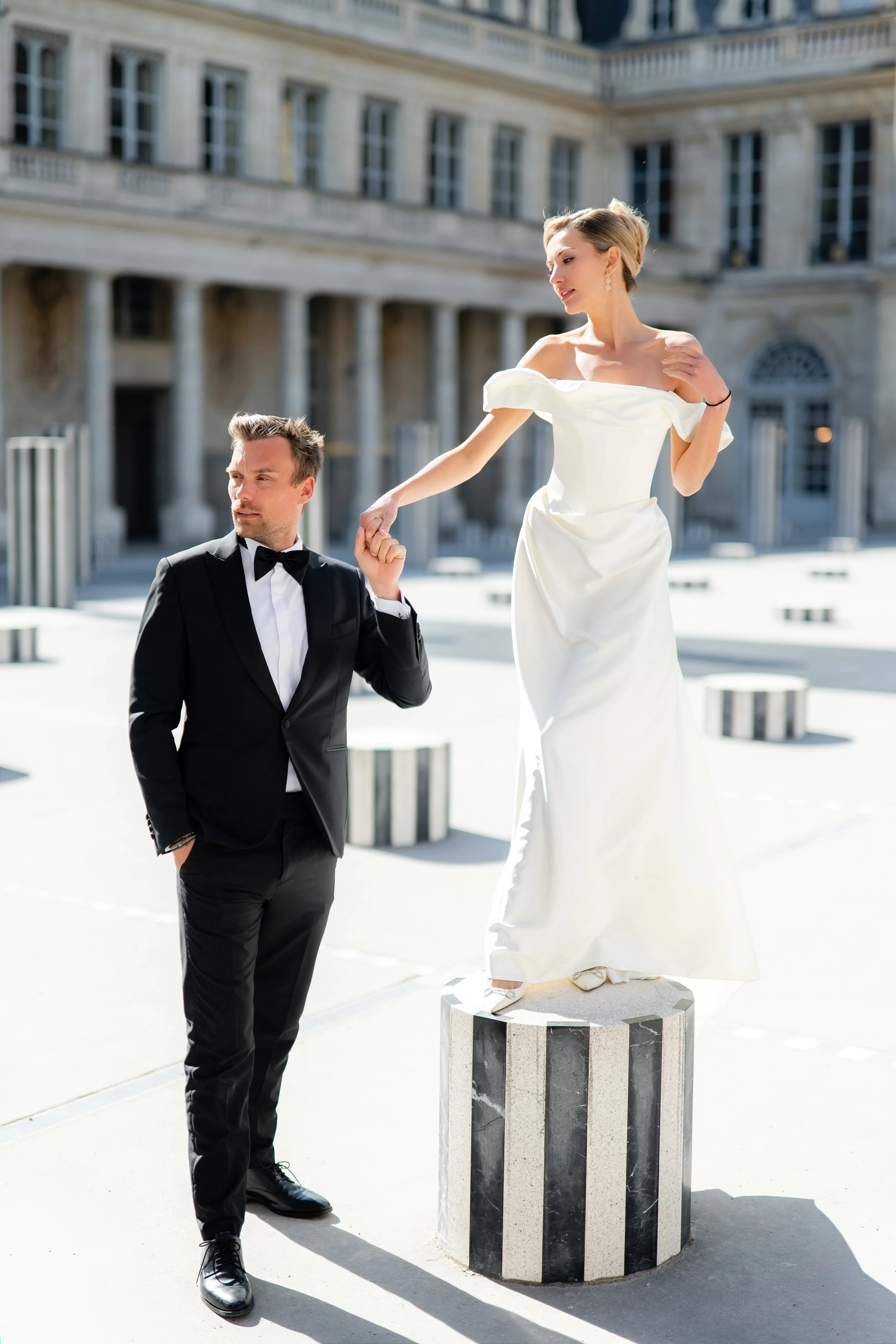 A man in a tuxedo kneeling and holding hands with a woman in a white gown, who is standing on a pedestal outdoors in front of a historic building.