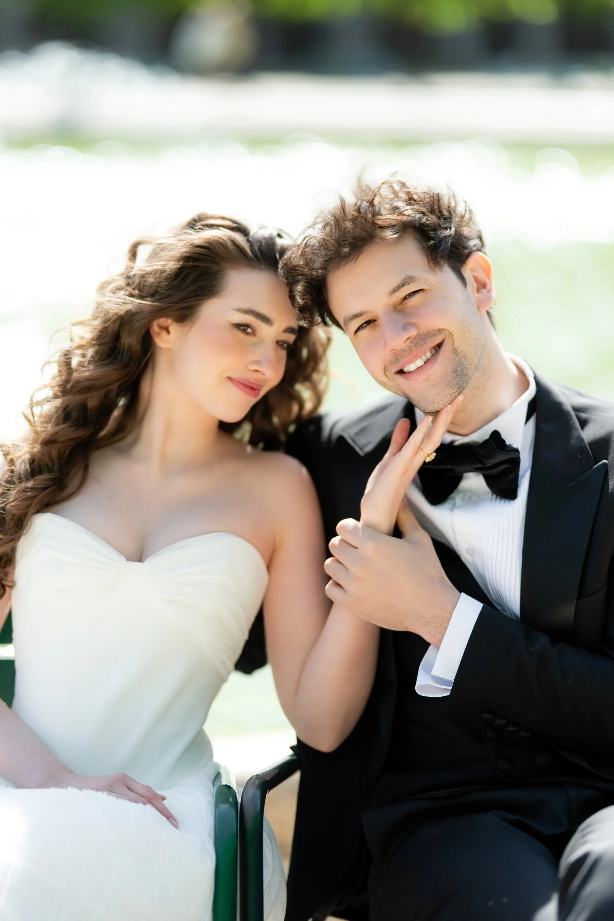 A young couple dressed in wedding attire sitting outdoors near a body of water, smiling and posing for a photo.