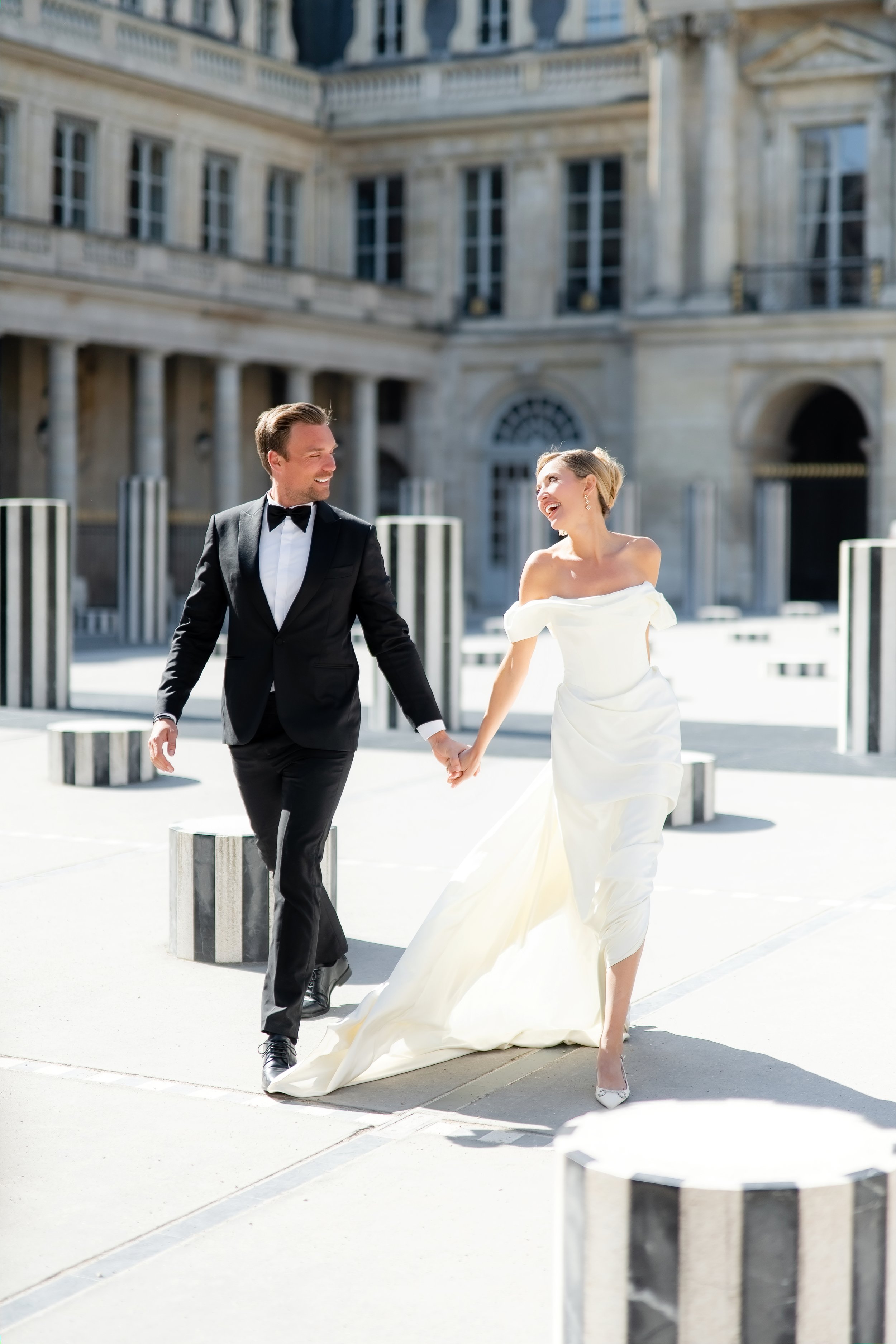 A smiling bride and groom holding hands and walking outdoors on a sunny day, with a historic stone building in the background.
